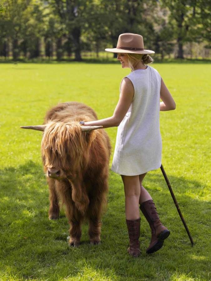 A woman in a sleeveless dress and hat stands on grass, holding the horn of a small, shaggy-haired cow