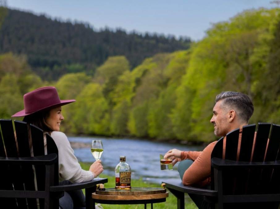 Two people sit in outdoor chairs by a river, enjoying drinks.