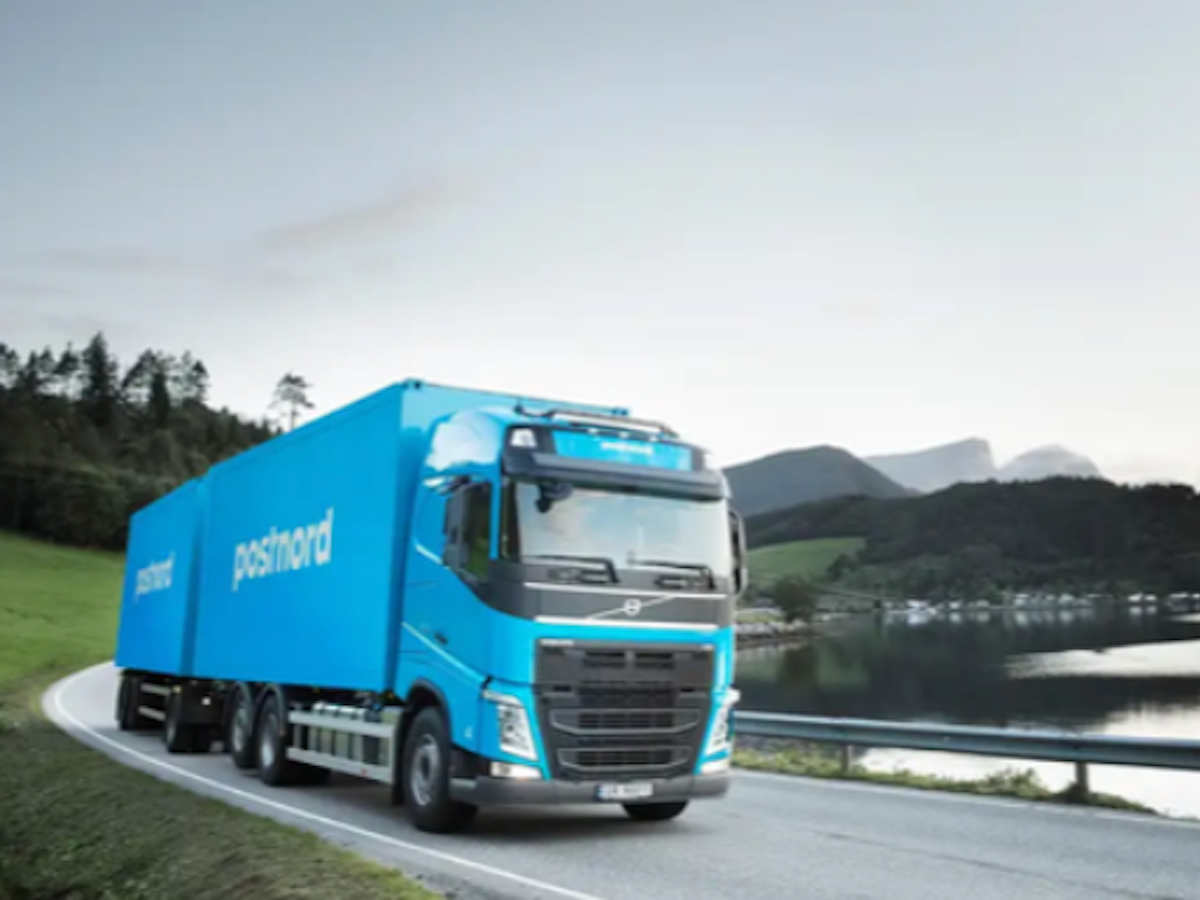 Blå PostNord-lastbil kör på landsväg bredvid en sjö med berg i bakgrunden.