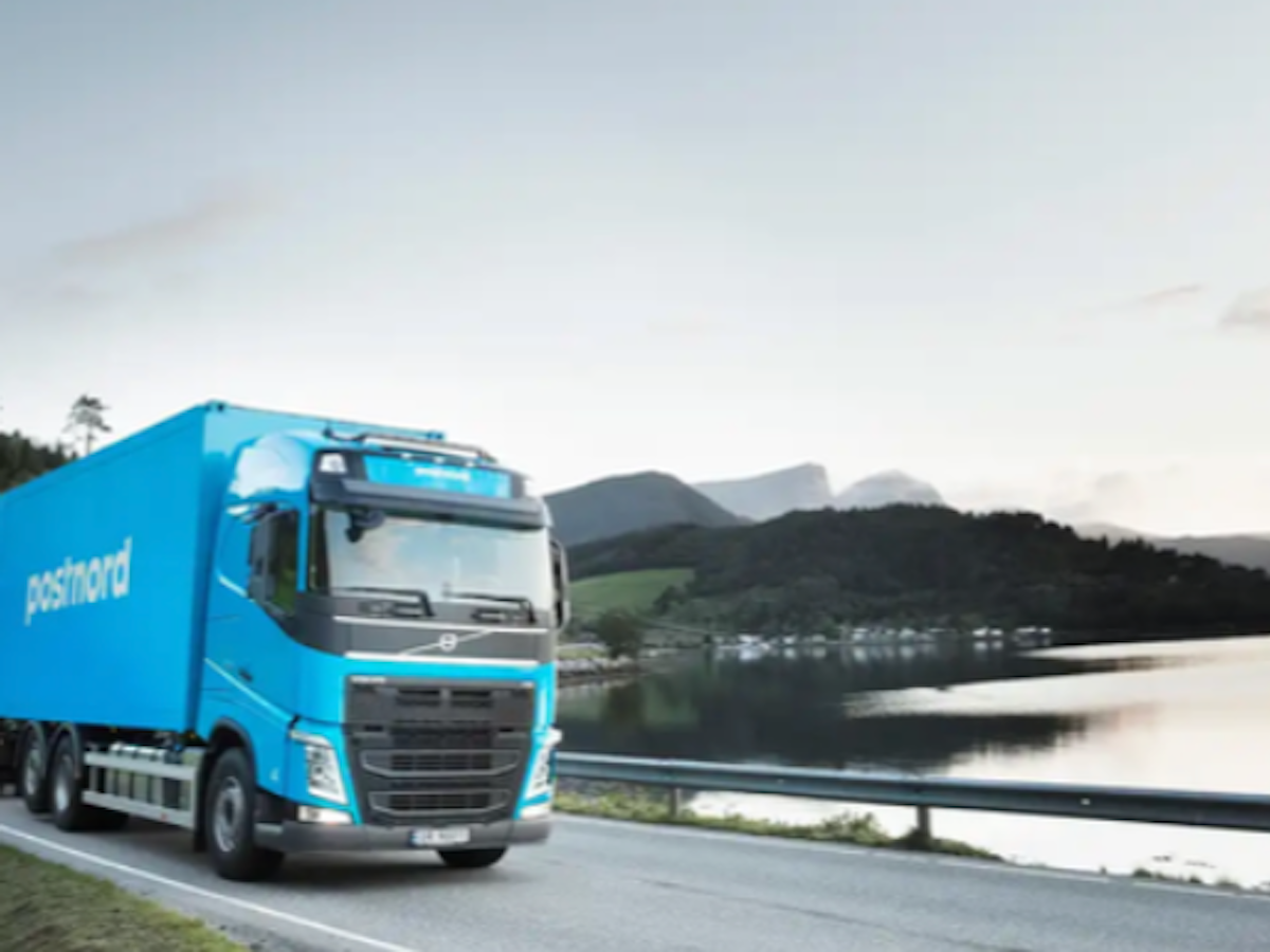 Blå PostNord-lastbil kör på landsväg bredvid en sjö med berg i bakgrunden.