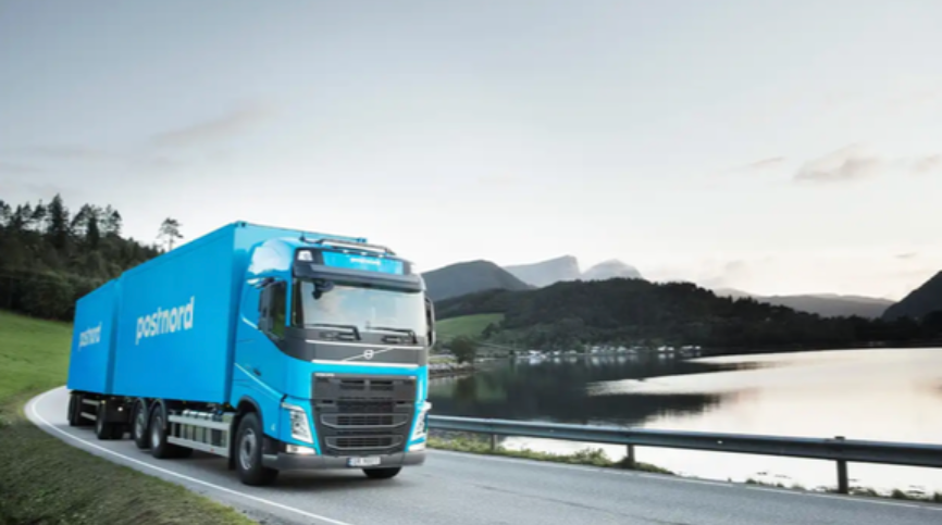 Blå PostNord-lastbil kör på landsväg bredvid en sjö med berg i bakgrunden.