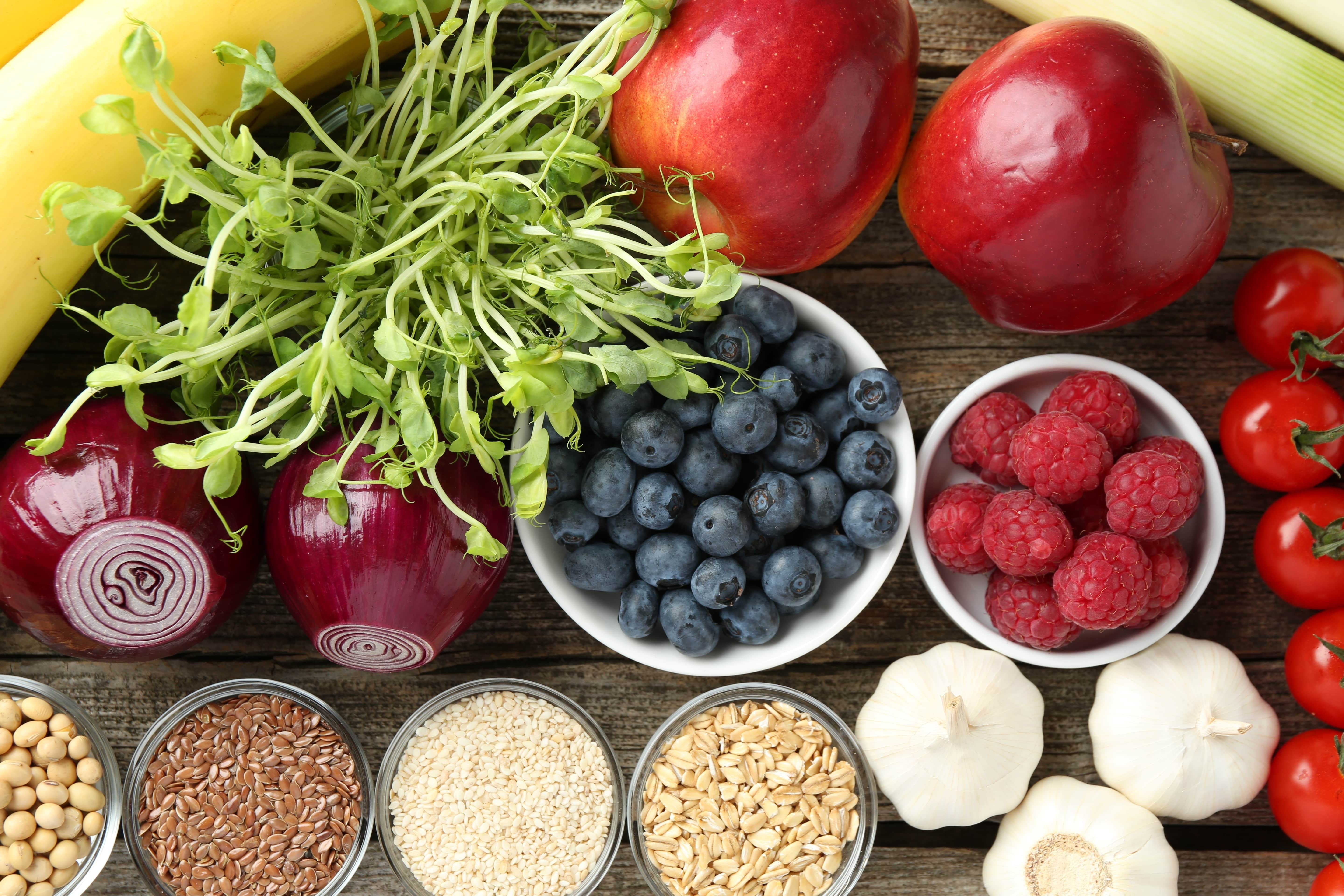 Fresh fruits, vegetables, grains and seeds including apples, berries, tomatoes, onions and garlic displayed on a wooden surface, representing a healthy balanced diet rich in plant-based nutrients