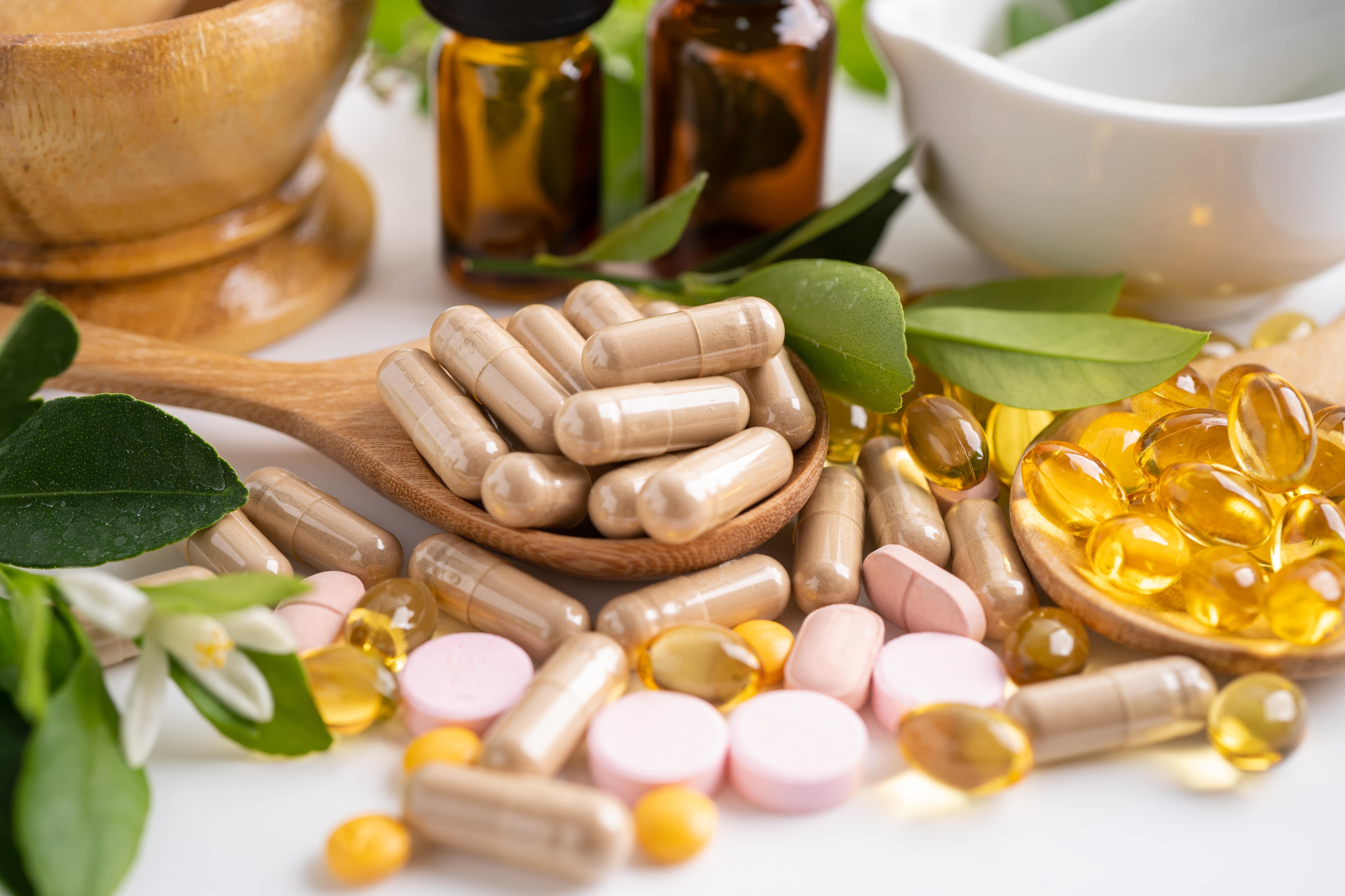 Assorted herbal supplements, vitamins and capsules on wooden spoons with natural leaves and glass bottles in the background