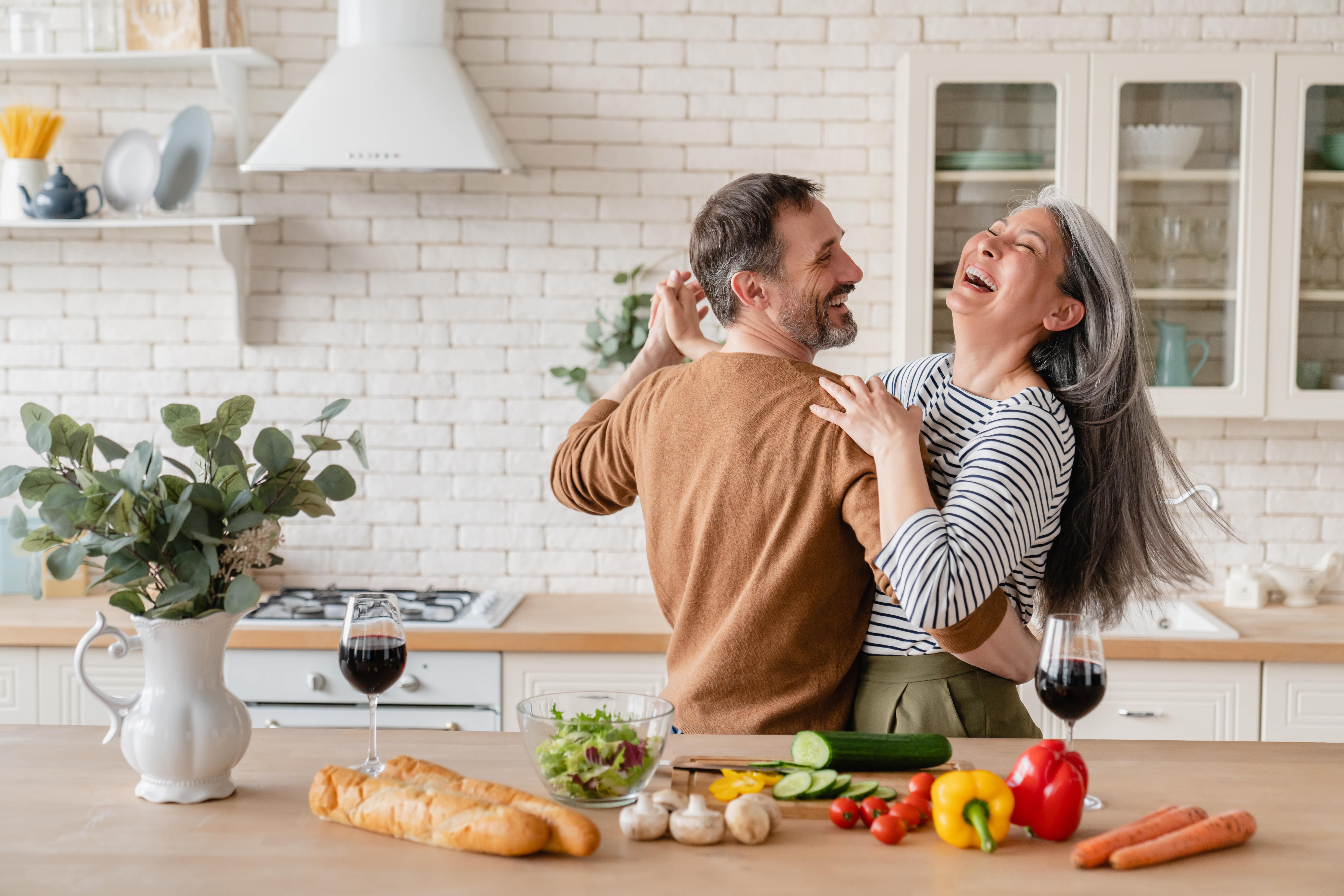 Happy middle-aged couple dancing together in a bright kitchen, surrounded by fresh vegetables, bread and wine, highlighting the joy of healthy living and cooking at home.