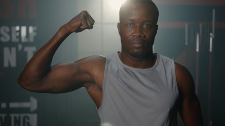 Confident man in gym flexing bicep in a sleeveless top, symbolising strength, fitness, and muscle training.