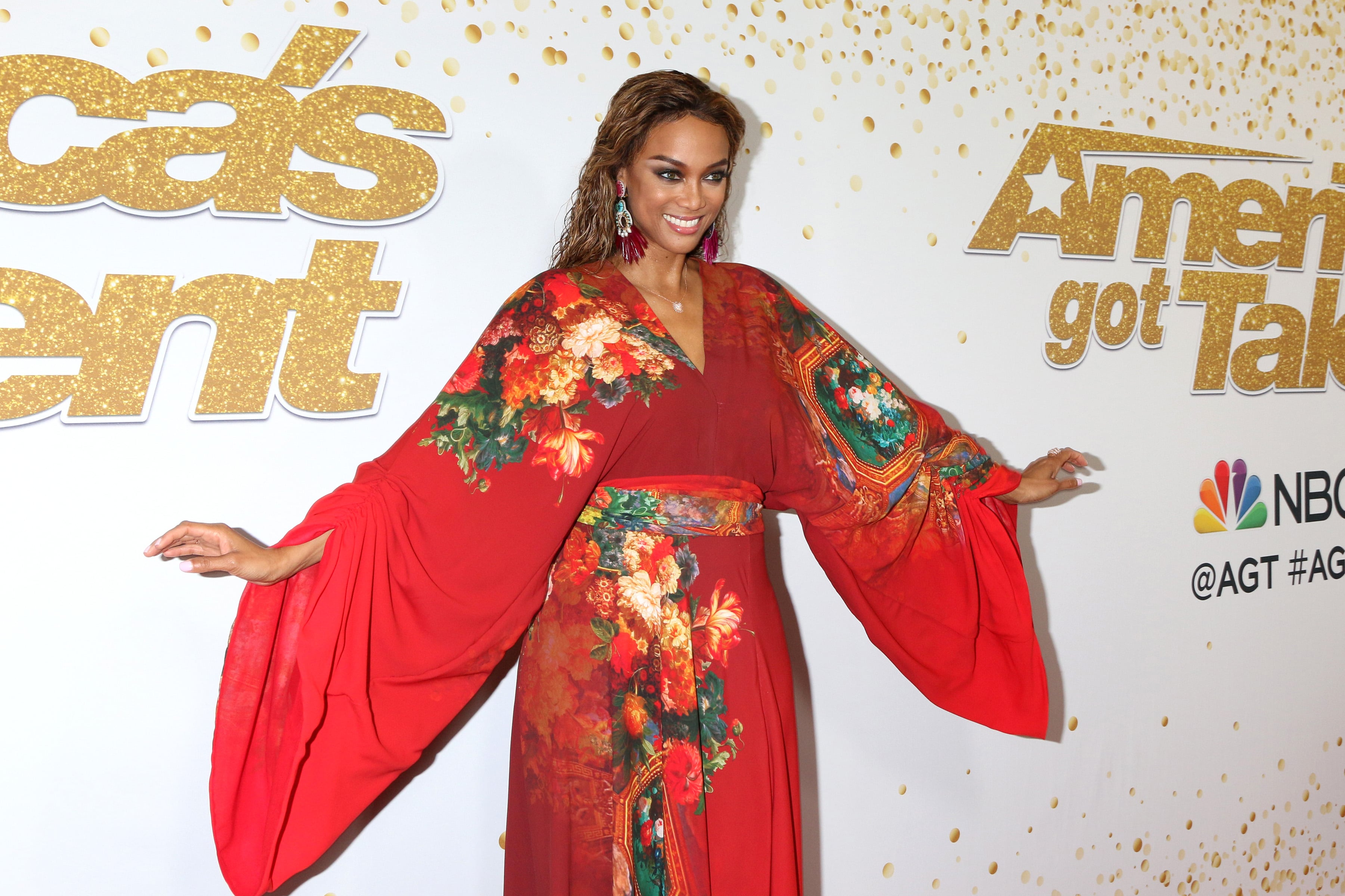 Tyra Banks posing in a red floral dress at an America's Got Talent event, representing high-profile celebrities who have opened up about managing IBS and digestive health issues.