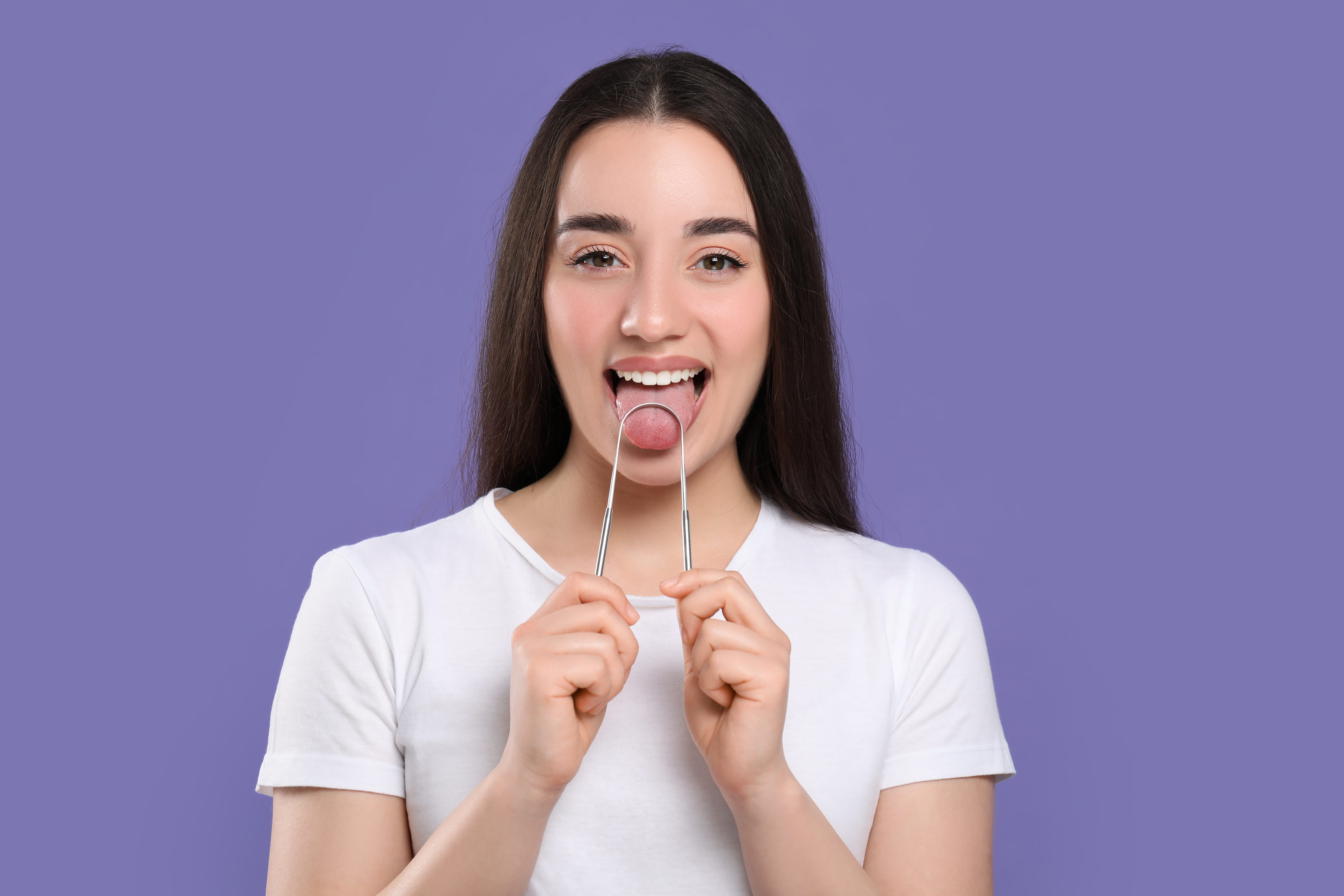 A young woman demonstrating the correct position of a metal tongue scraper on her tongue, visually representing the first step in an effective oral hygiene routine for fresher breath and better dental health.
