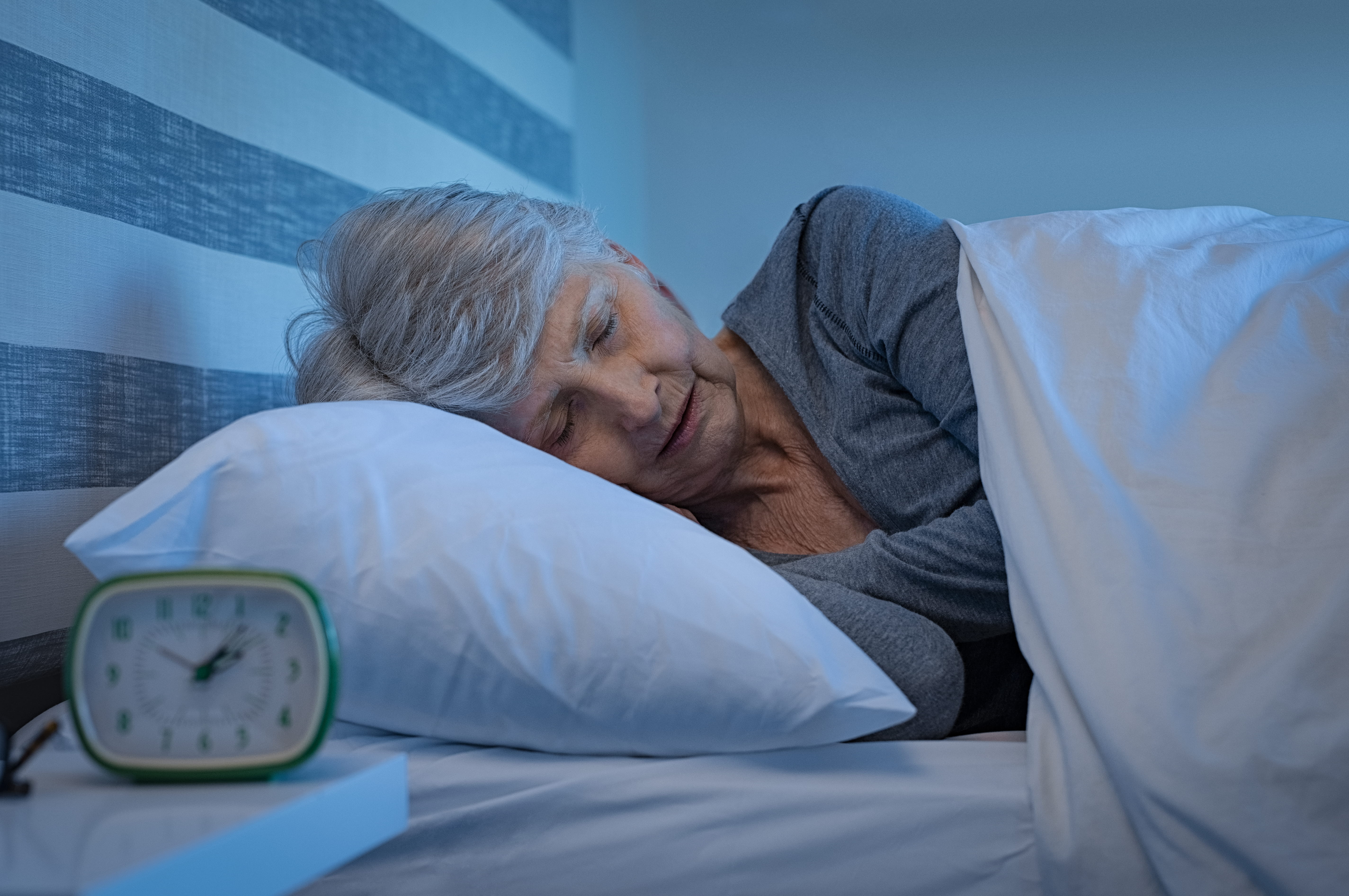 A senior woman sleeping peacefully in a dark bedroom with an alarm clock on her bedside table, visually representing the deep, restorative sleep necessary for the brain to clear harmful toxins and lower the risk of Alzheimer's disease.