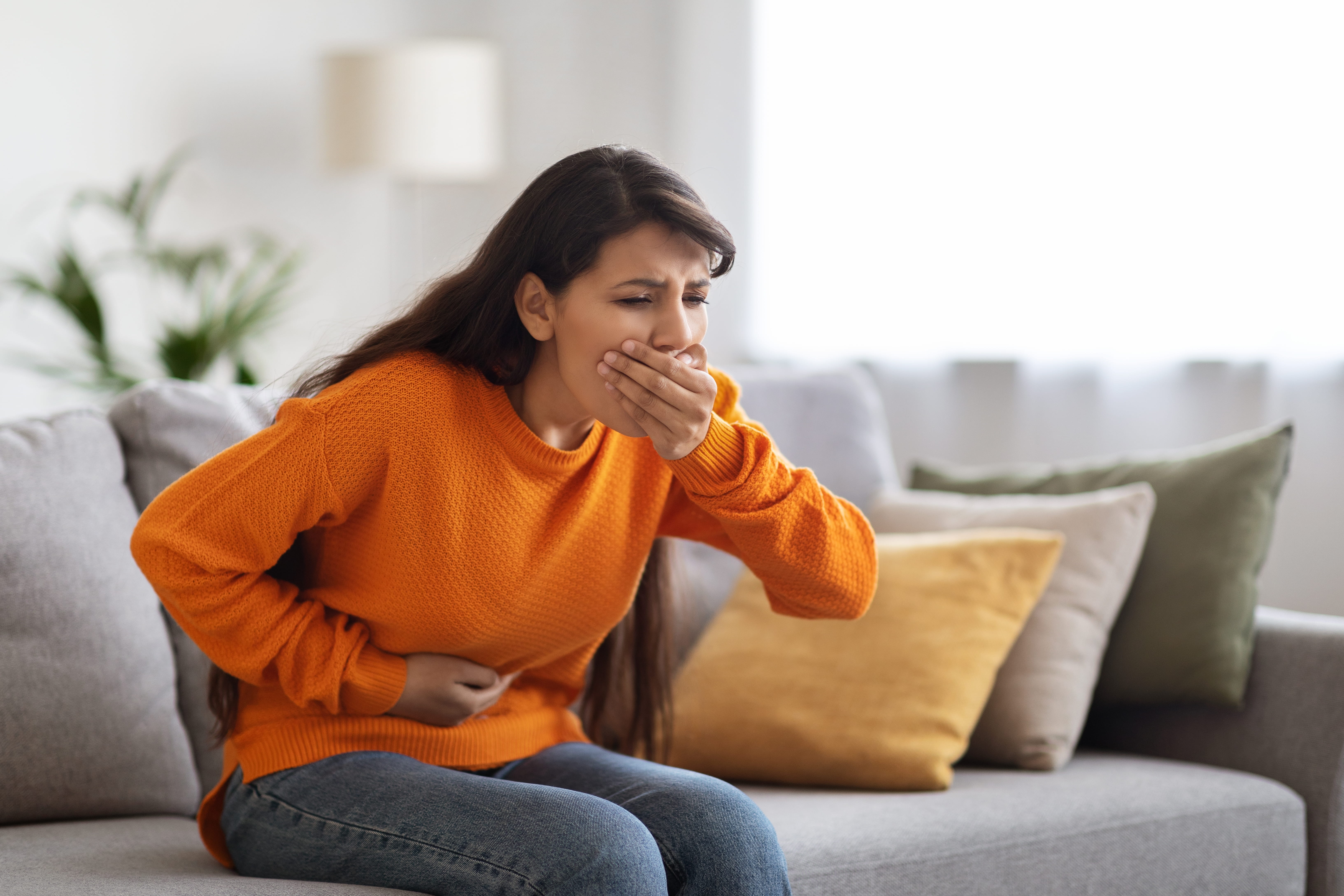Woman on a sofa with one hand over her mouth and the other on her stomach, looking nauseous