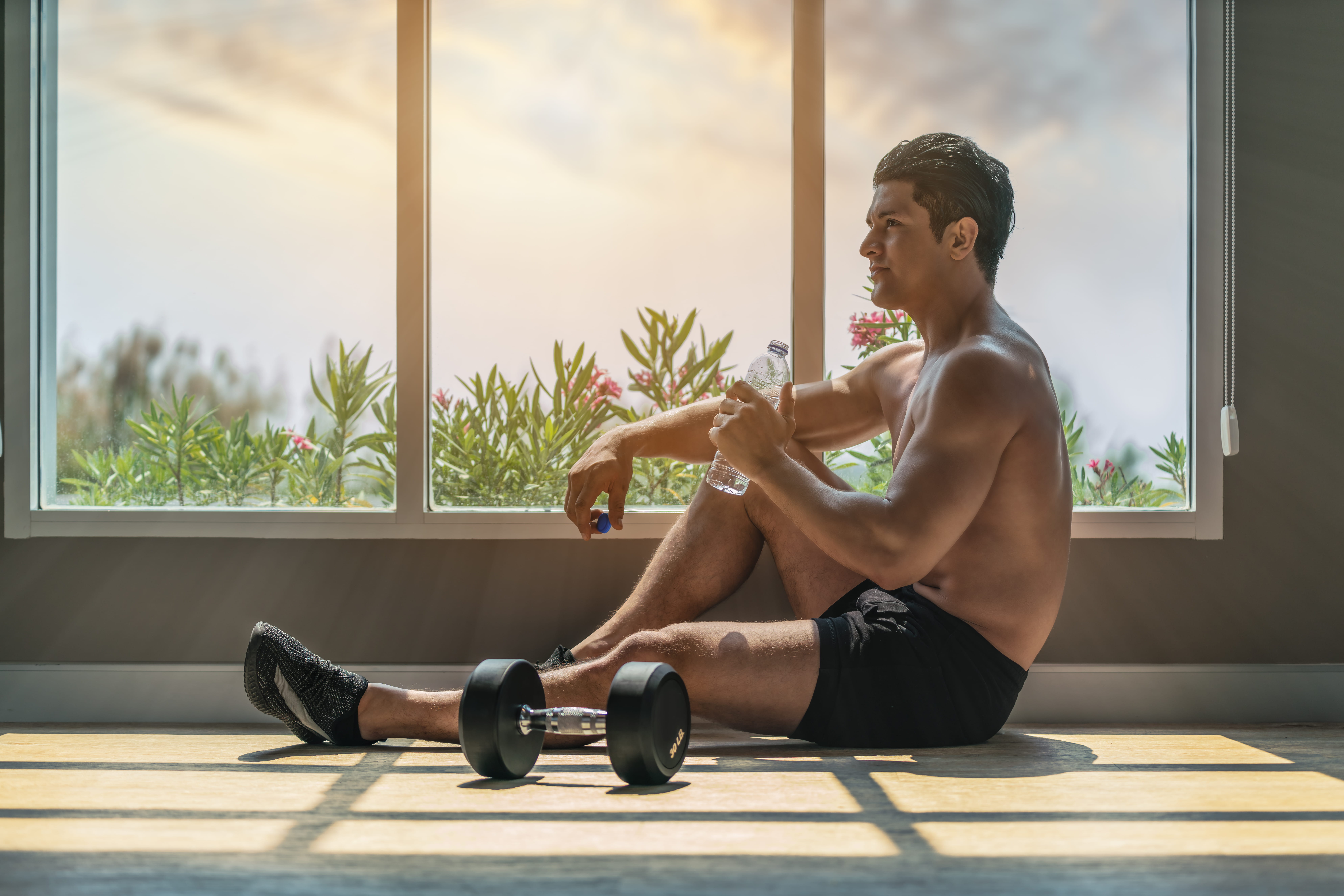 Man resting after a workout by a window, holding a shaker bottle with a dumbbell beside him