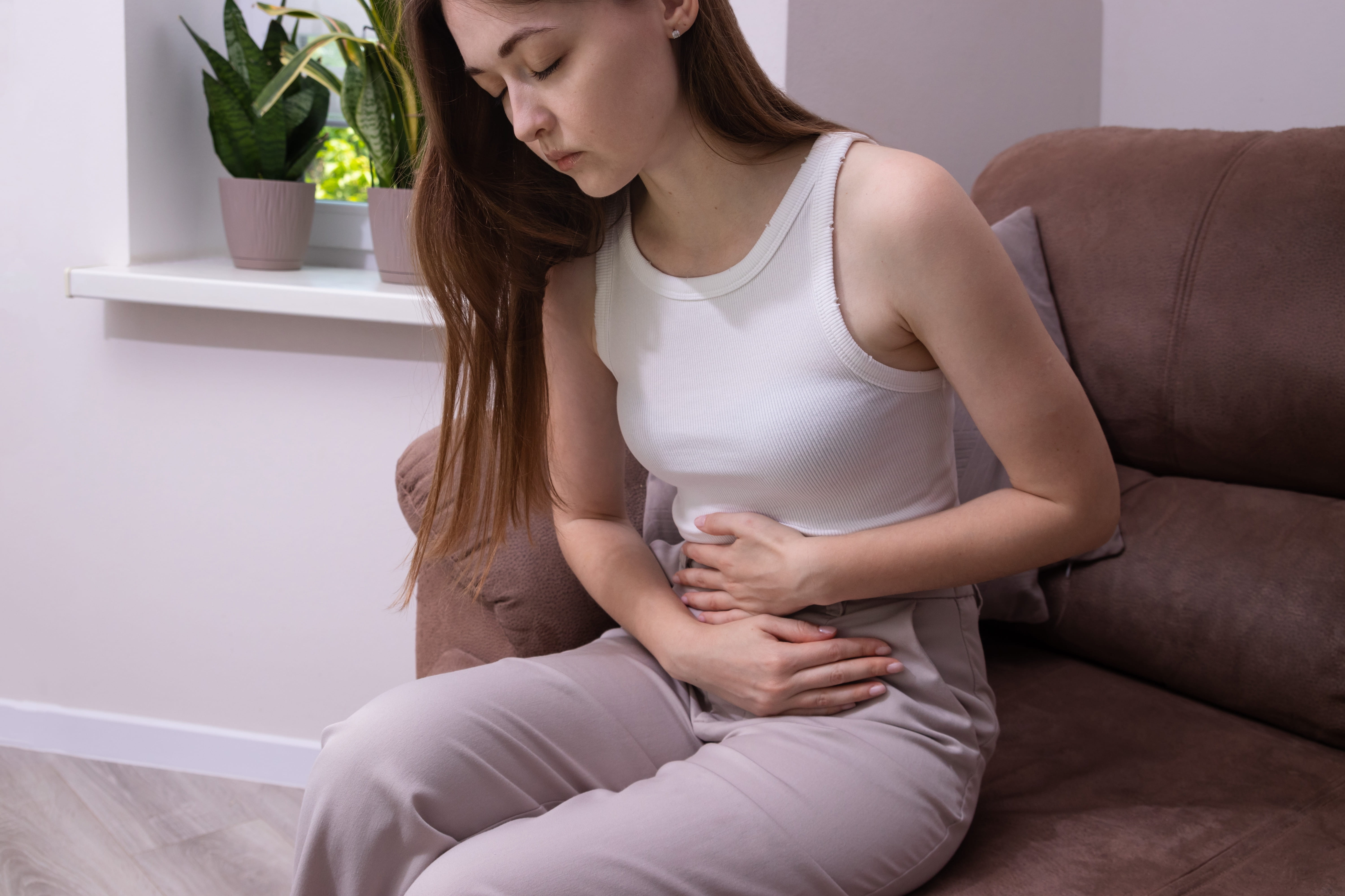 Woman sitting on a sofa with both hands resting on her stomach, looking uncomfortable with bloating