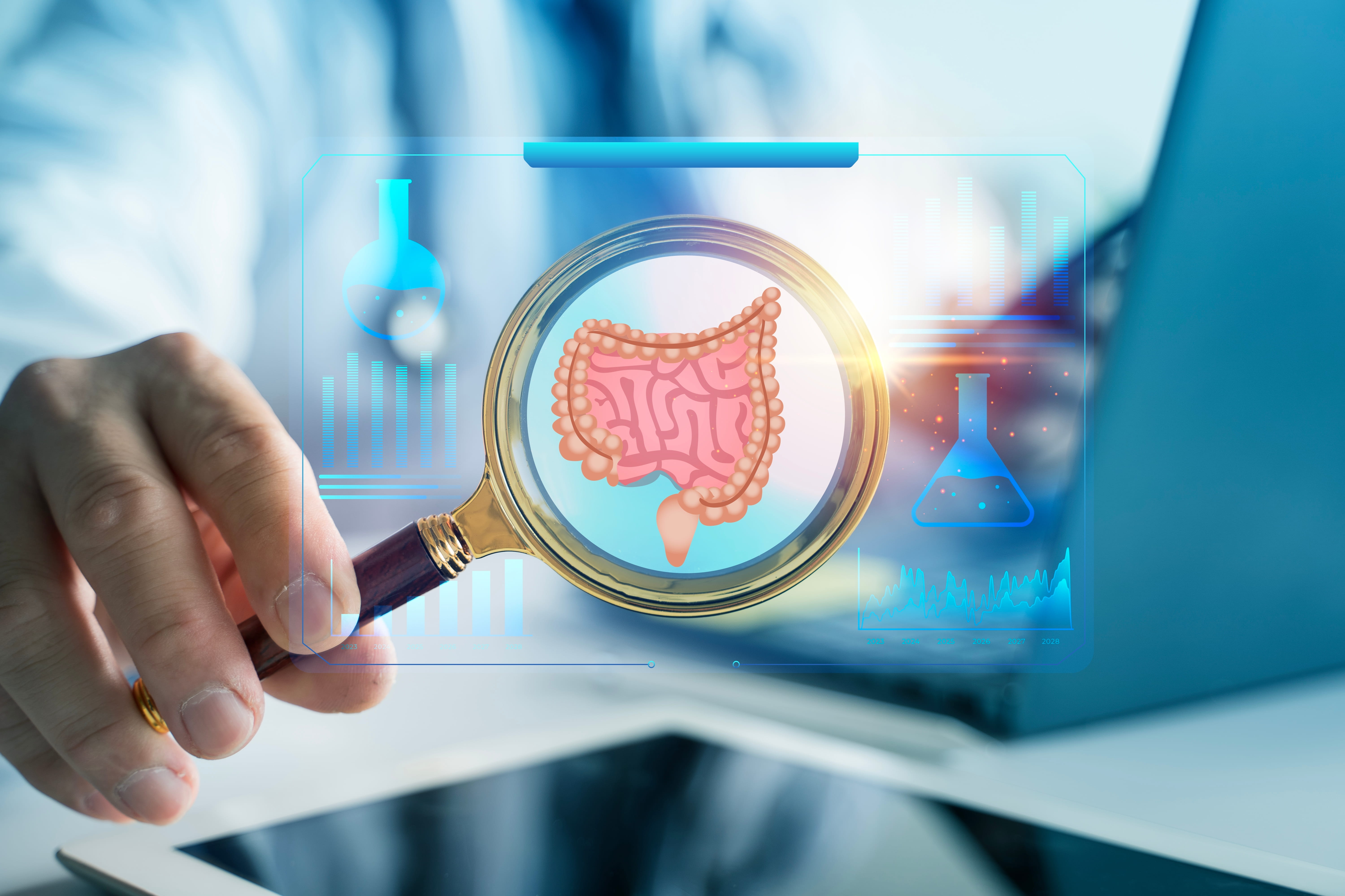 Hand holding a magnifying glass over a glowing digital illustration of the gut, with a laptop behind