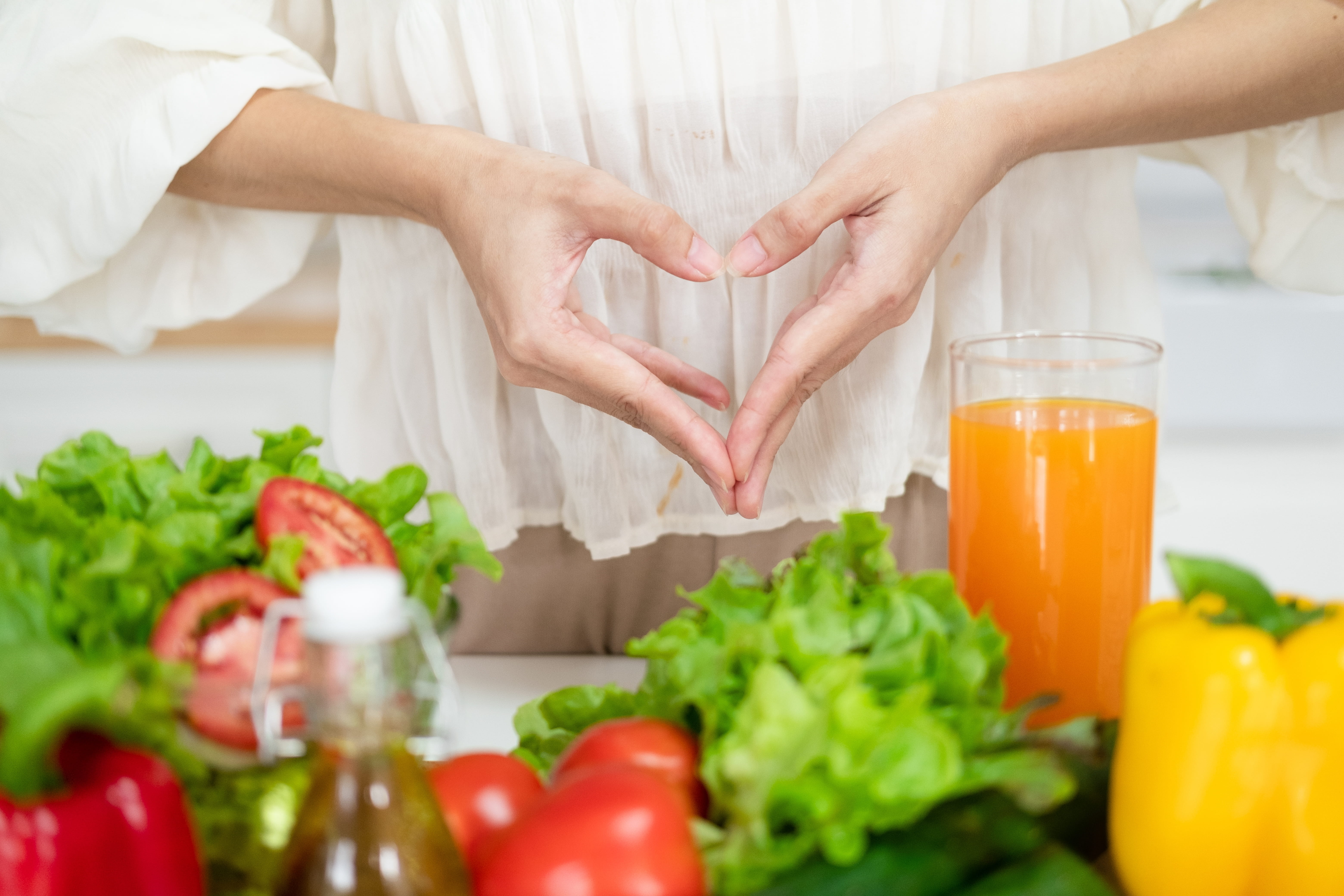 Hands forming a heart shape over the stomach with fresh vegetables and a glass of juice in front