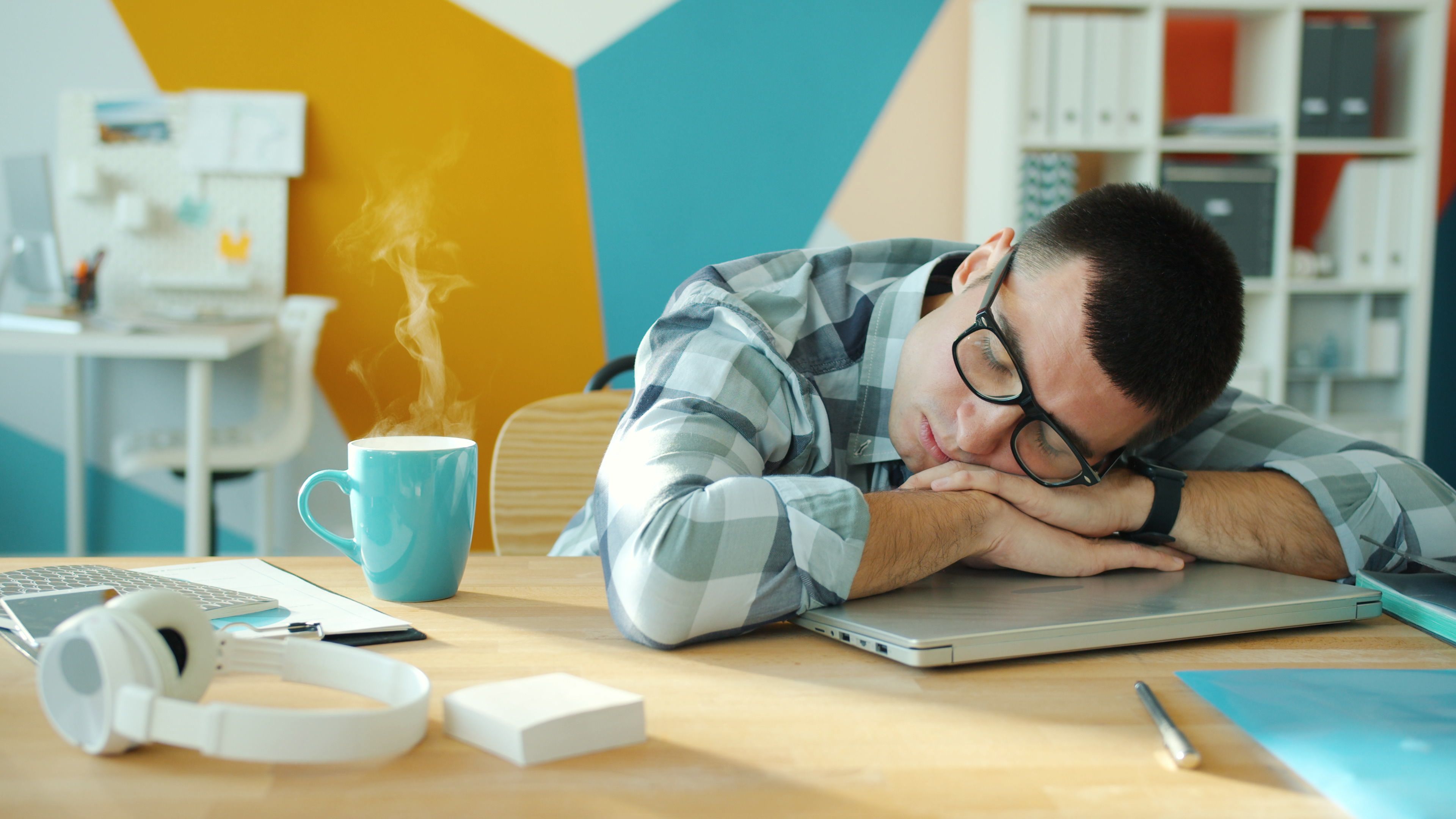 Tired man slumped with his head resting on a laptop at a desk, a coffee mug beside him
