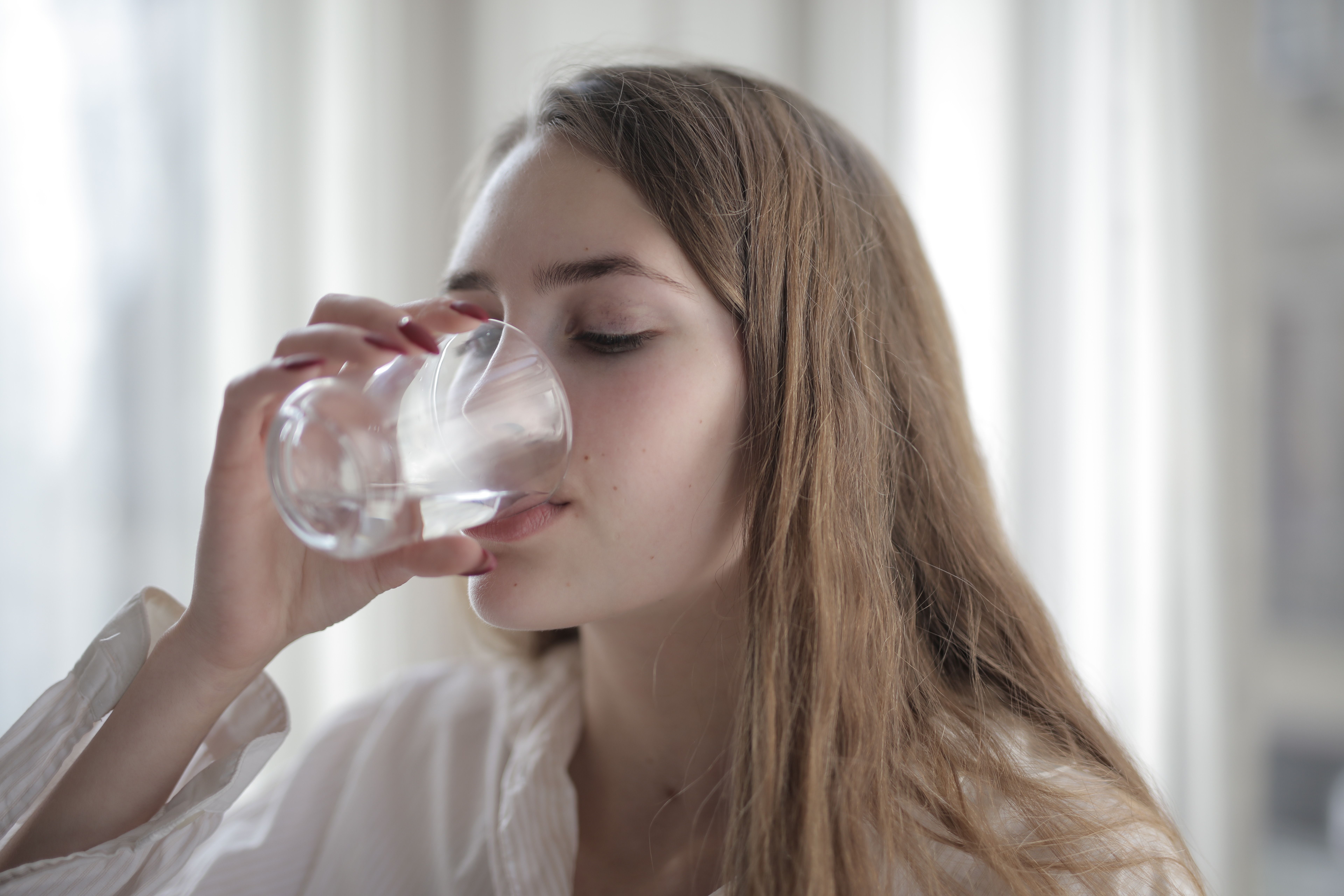 Woman drinking a glass of water, taking a capsule with her eyes closed by a bright window