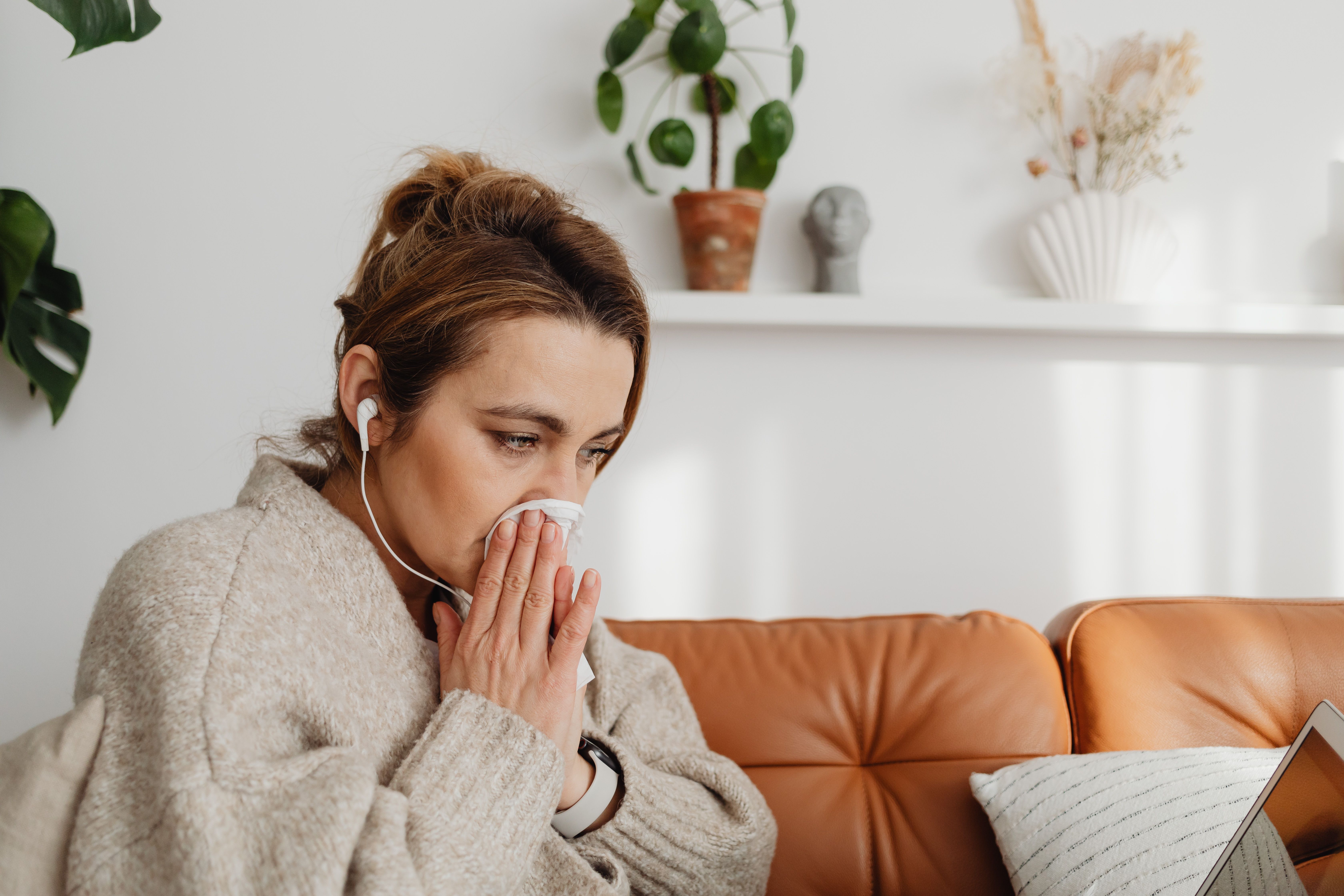 Woman wrapped in a blanket on a sofa, looking unwell and blowing her nose or feeling cold
