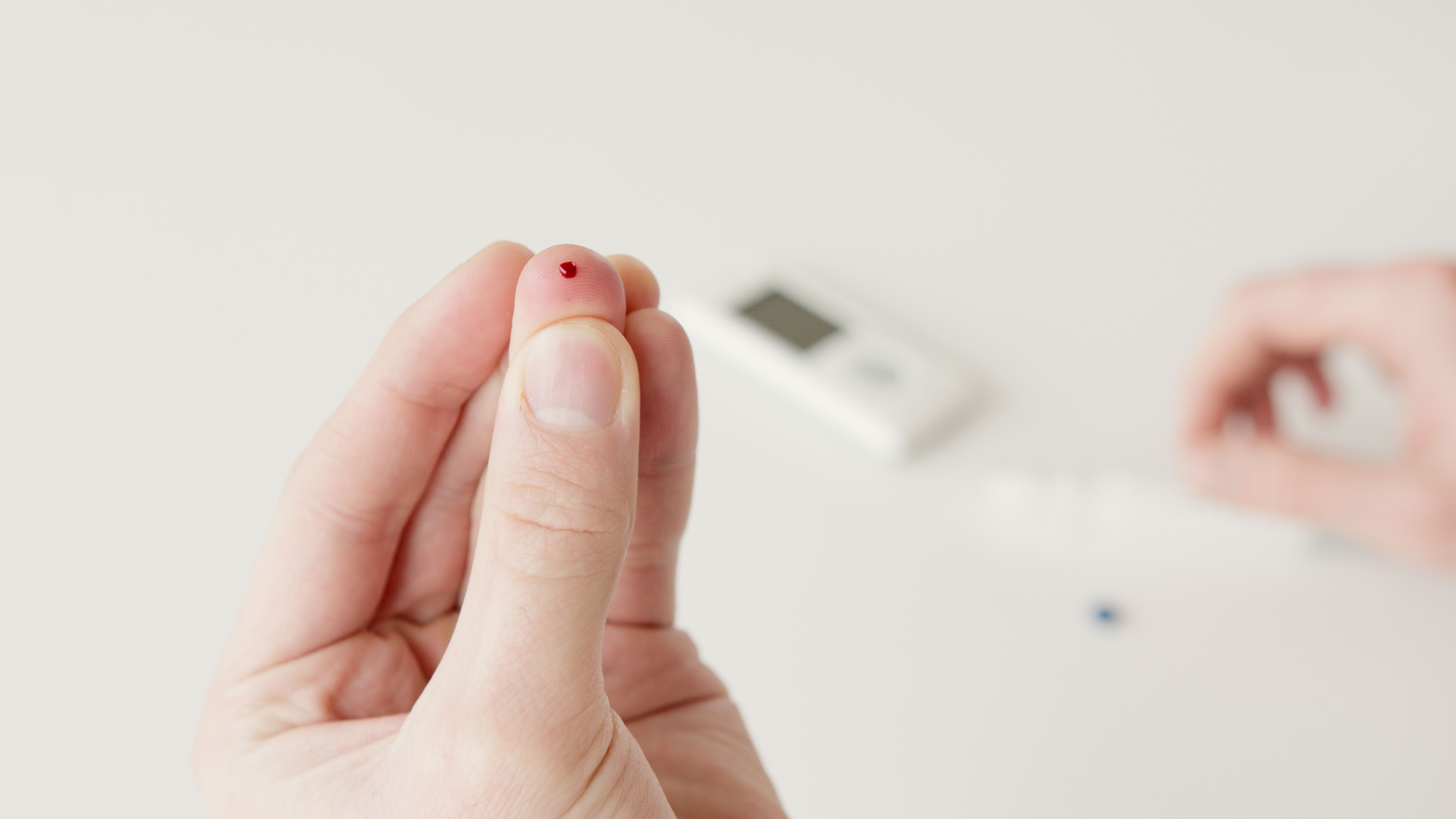 Finger with a drop of blood held up for a blood sugar test, glucose meter blurred behind