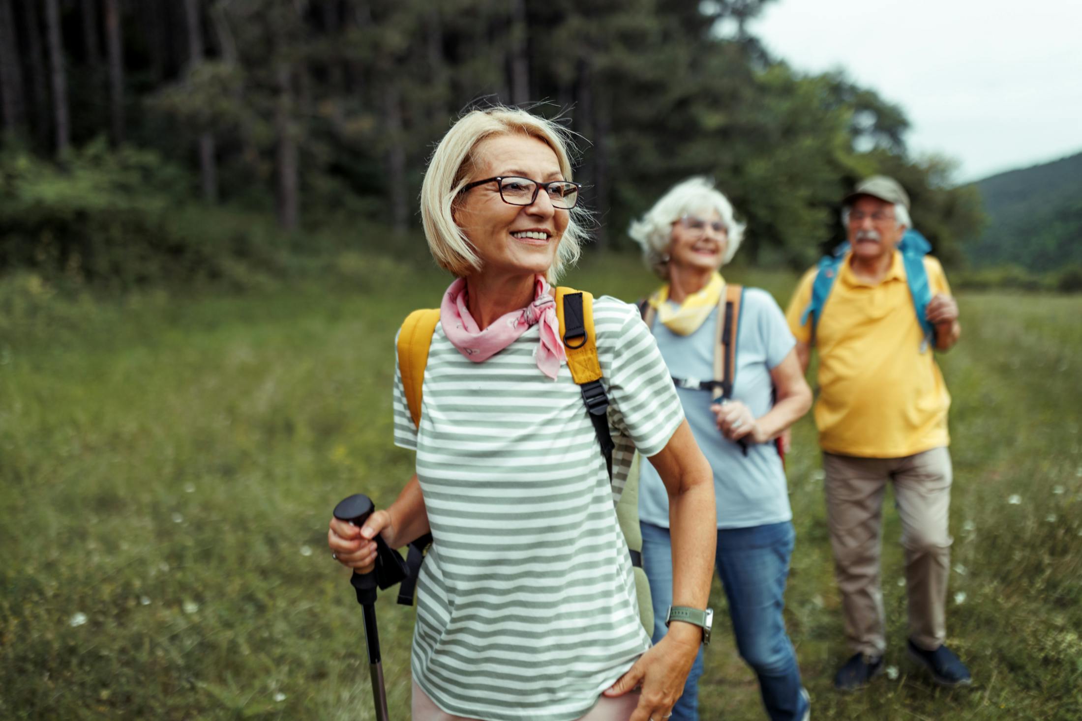 2 older women and an older man are hiking in a beautiful meadow lined with trees. They are smiling and showing the benefits of having no foot or bunion pain.
