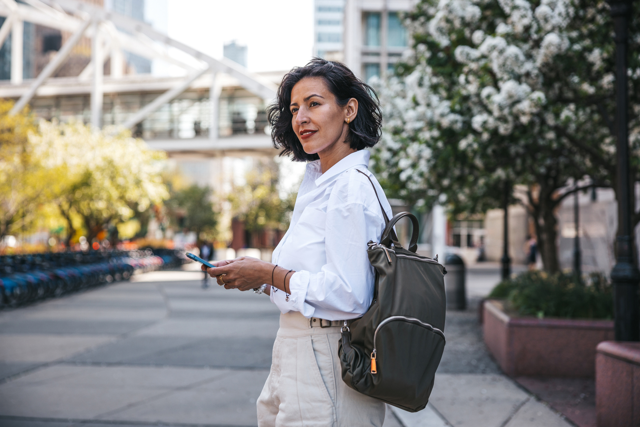 Mature woman standing outdoors in an urban setting with a backpack and smartphone.