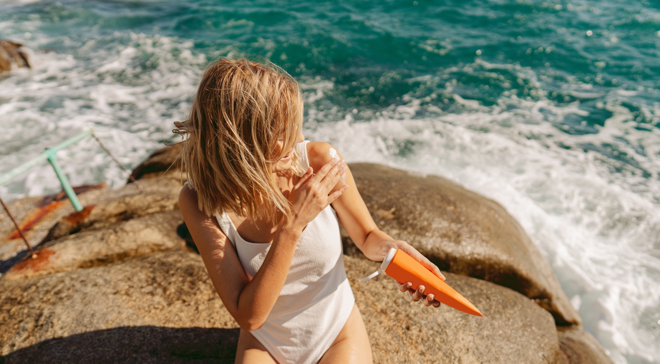 Woman applying sunscreen to her shoulder while sitting by the ocean.