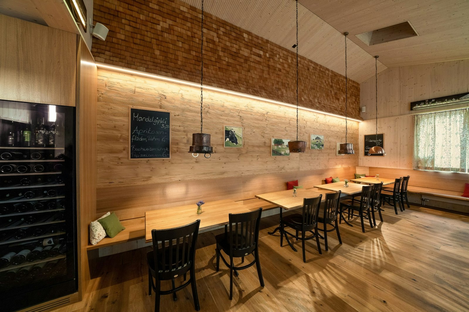 Ein gemütliches Restaurant mit Holzwänden, Tischen und Stühlen, Hängelampen, einem Weinkühler, einer Speisekarte auf Kreidetafeln und gerahmten Fotos.