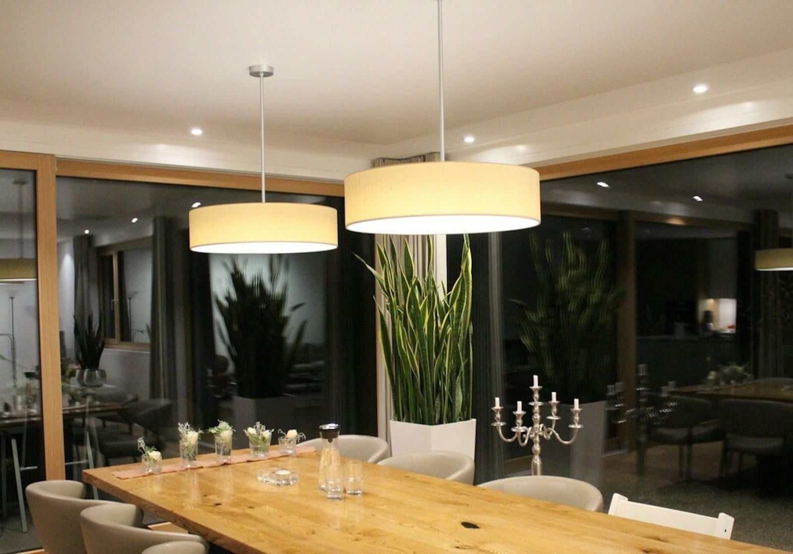 Modernes Esszimmer mit Holztisch, weissen Stühlen, zwei grossen Pendelleuchten, Topfpflanzen, Kandelabern und grossen Glasfenstern.