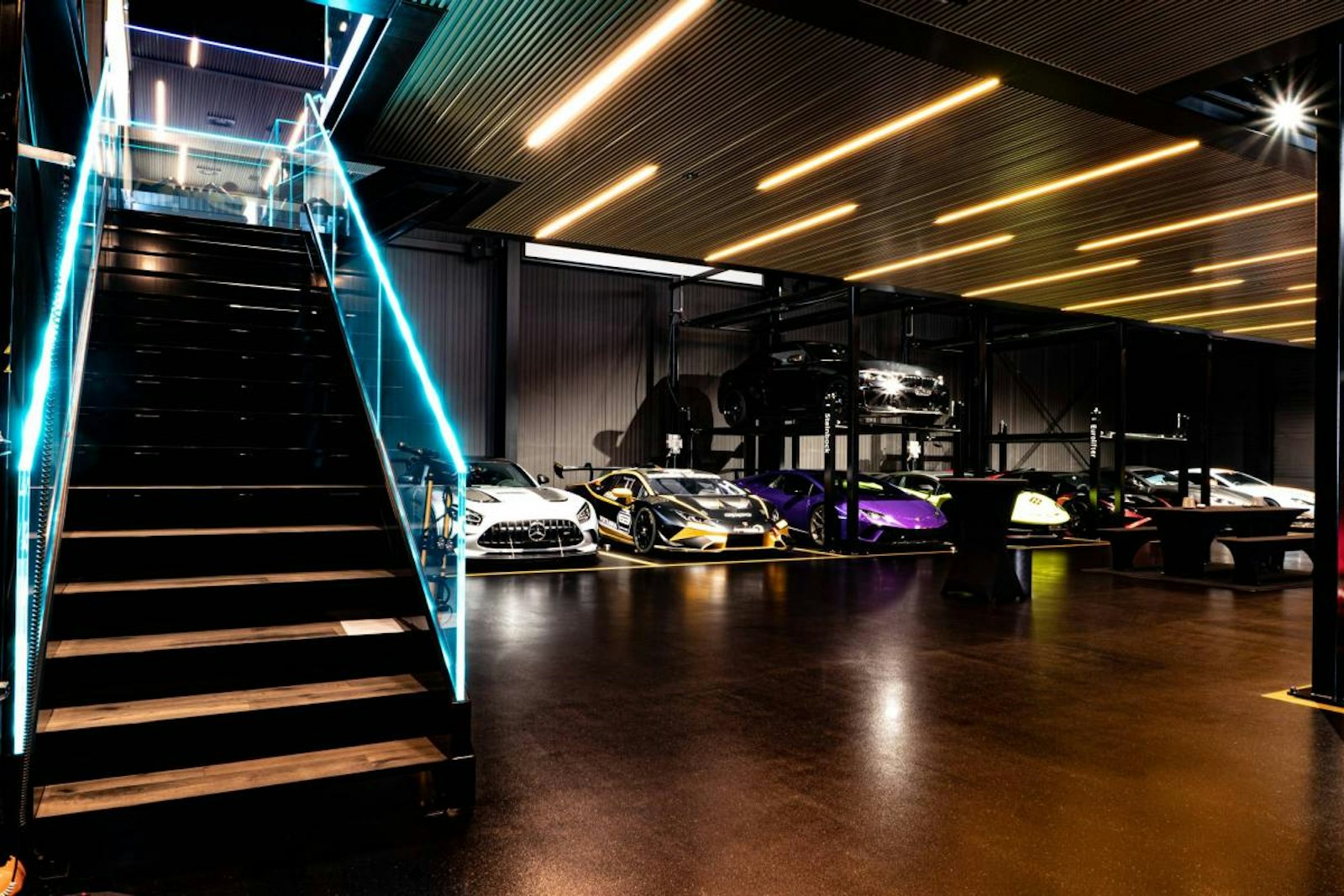 Eine moderne überdachte Garage beherbergt Luxussportwagen auf zwei Ebenen neben einer Treppe mit blaür Beleuchtung und einem polierten Boden.