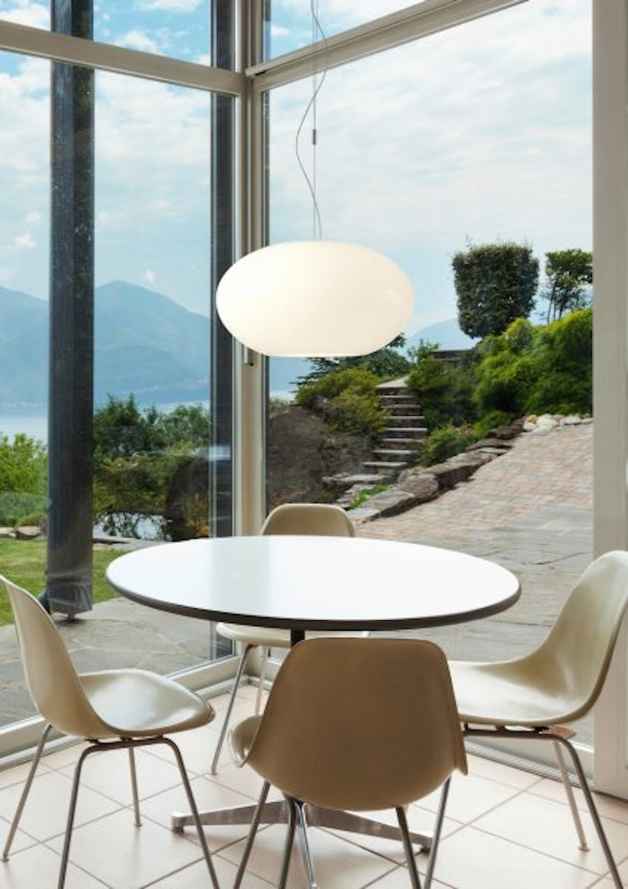 Ein runder weisser Tisch mit vier beigen Stühlen steht an grossen Fenstern mit Blick auf den Garten und die Berge, darüber eine moderne Pendelleuchte.