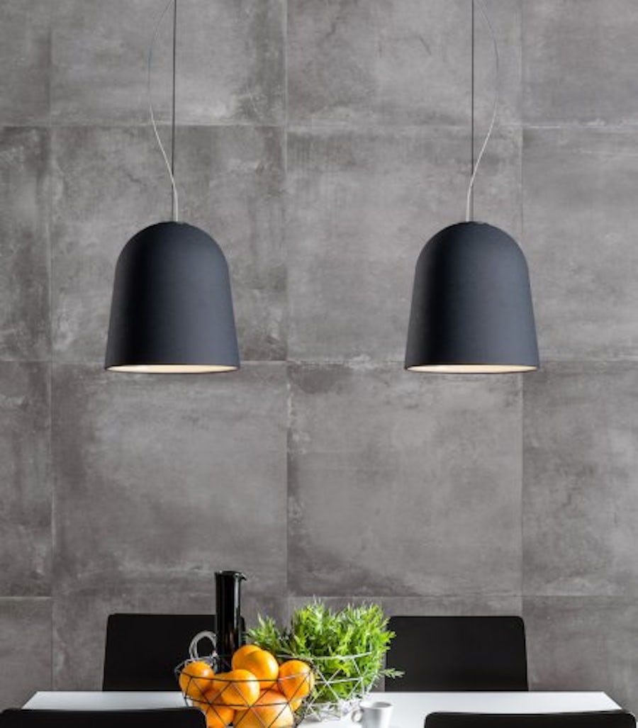 Schwarze Pendelleuchten hängen über einem weissen Tisch mit einem Obstkorb, einem schwarzen Krug, einem grünen Blumentopf, zwei schwarzen Stühlen und einer graün Kachelwand.