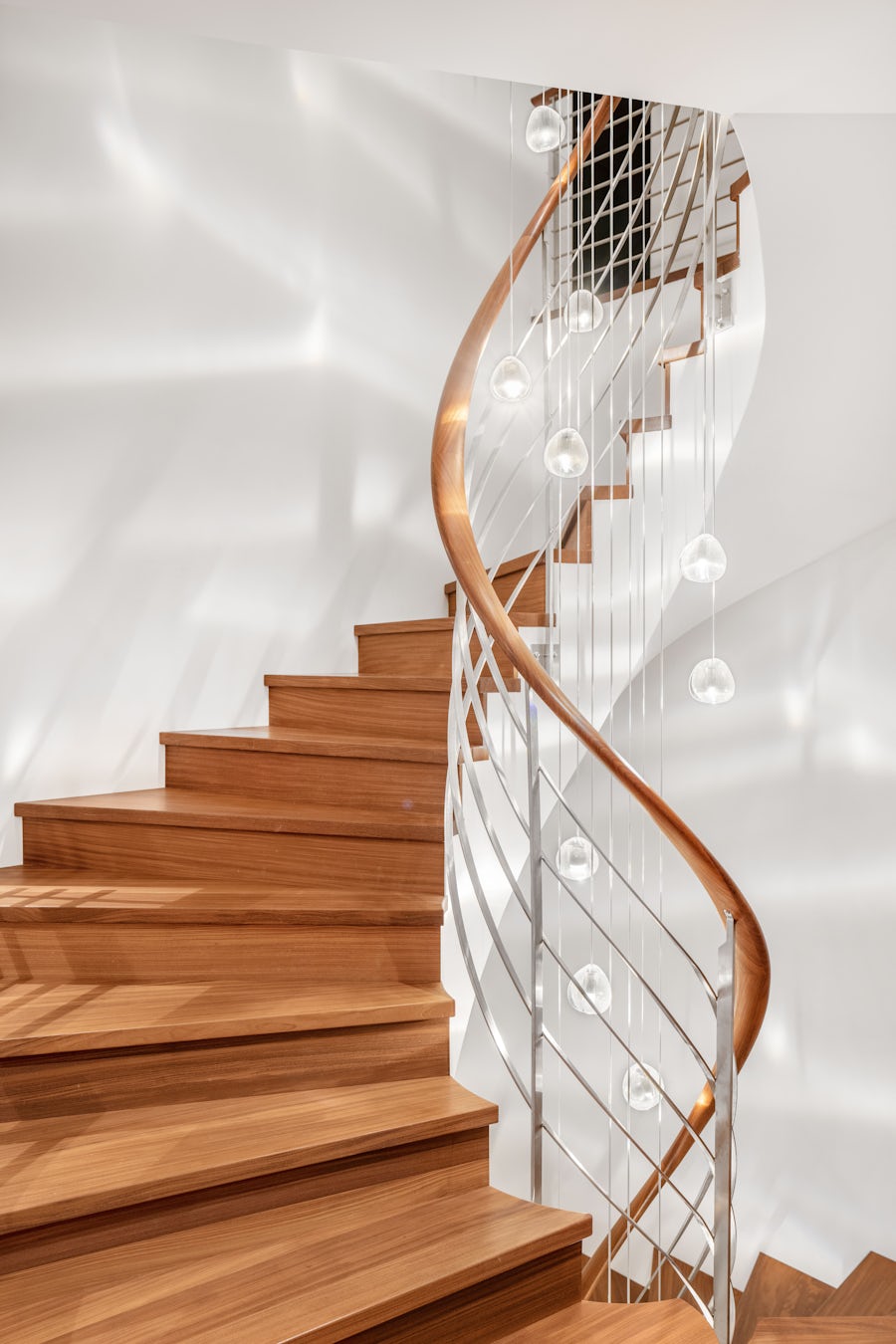 Eine geschwungene Holztreppe mit einem eleganten silbernen Geländer und Pendelleuchten an einer glatten weissen Wand.