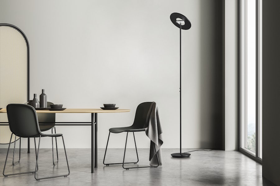 Minimalistischer Essbereich: zwei Stühle, Holztisch mit Geschirr, graü Decke auf einem Stuhl, Stehlampe, grosse Fenster mit Tageslicht.