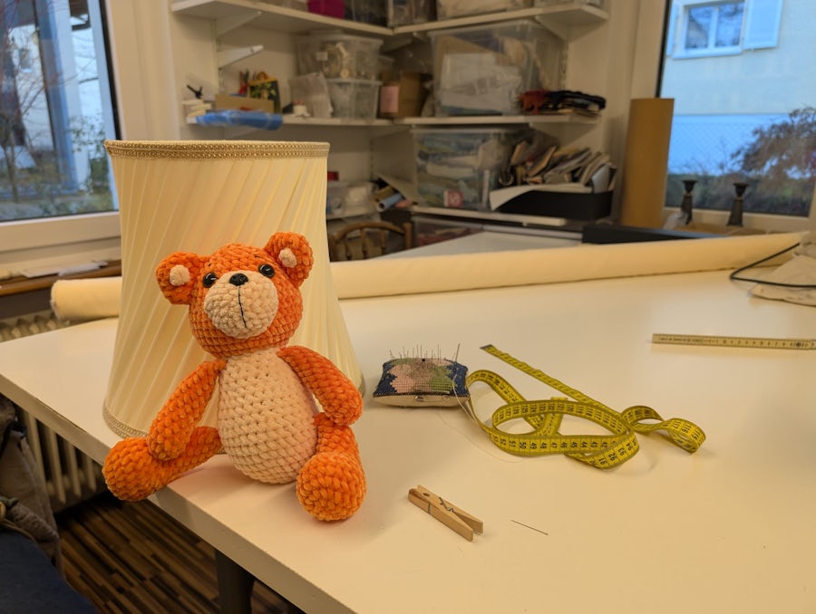 Orangefarbener gehäkelter Teddybär auf einem weissen Tisch mit Nähwerkzeug, Lampenschirm und Nadelkissen in einem hellen Bastelzimmer.