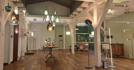 Ein geräumiger Ausstellungsraum mit Holzböden und sichtbaren Balken bietet moderne Steh- und Pendelleuchten in einem hellen, zeitgenössischen Raum.