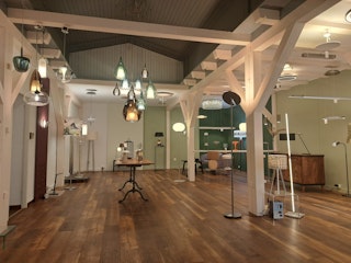 Ein geräumiger Ausstellungsraum mit Holzböden und sichtbaren Balken bietet moderne Steh- und Pendelleuchten in einem hellen, zeitgenössischen Raum.