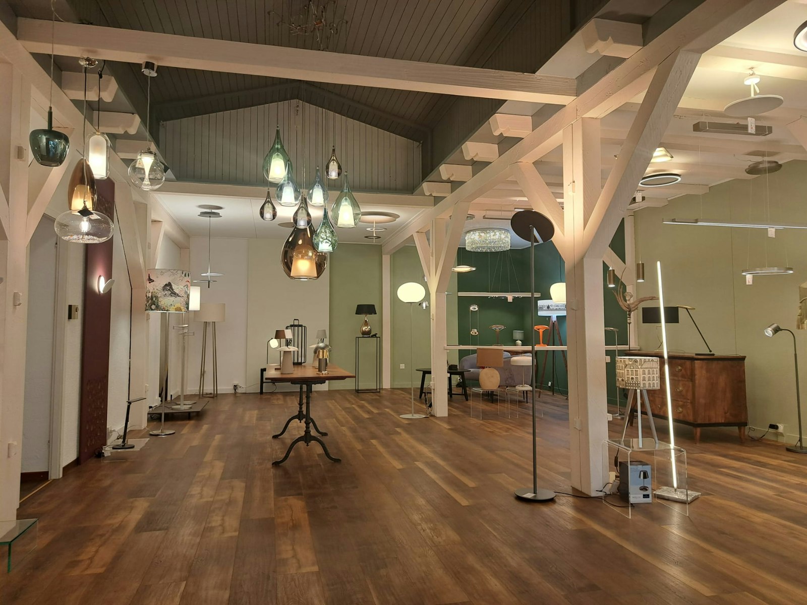 Ein geräumiger Ausstellungsraum mit Holzböden und sichtbaren Balken bietet moderne Steh- und Pendelleuchten in einem hellen, zeitgenössischen Raum.