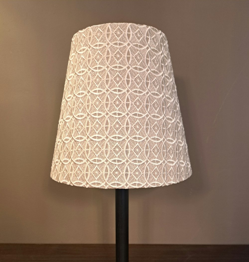 Ein beiger Lampenschirm mit geometrischem Spitzenmuster sitzt auf einem schwarzen Lampenfuss vor einer schlichten braunen Wand.