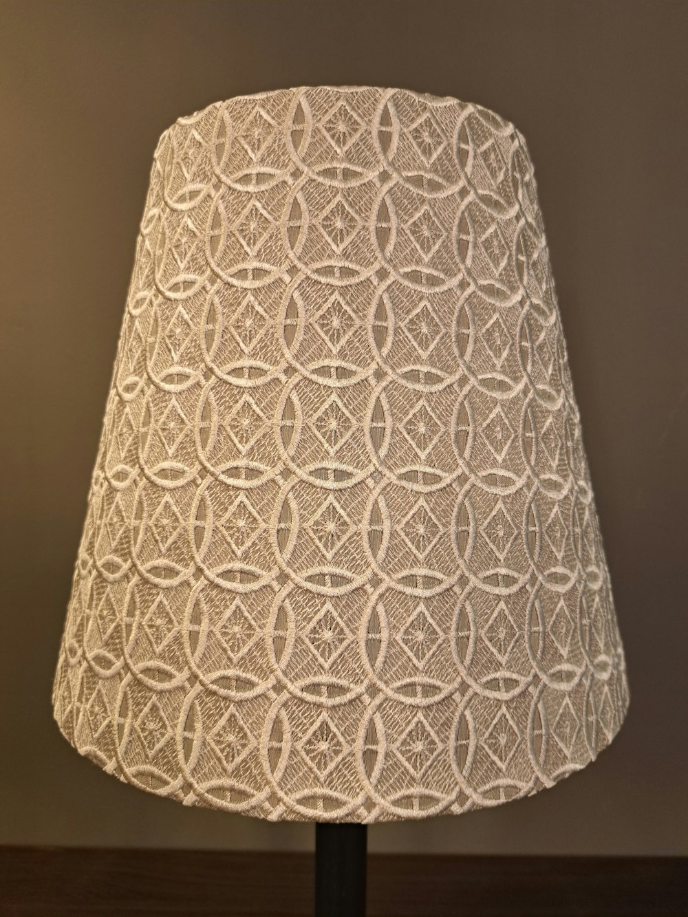 Nahaufnahme eines beigen Lampenschirms mit einem komplizierten geometrischen Muster aus Kreisen und Rauten vor einem schlichten braunen Hintergrund.