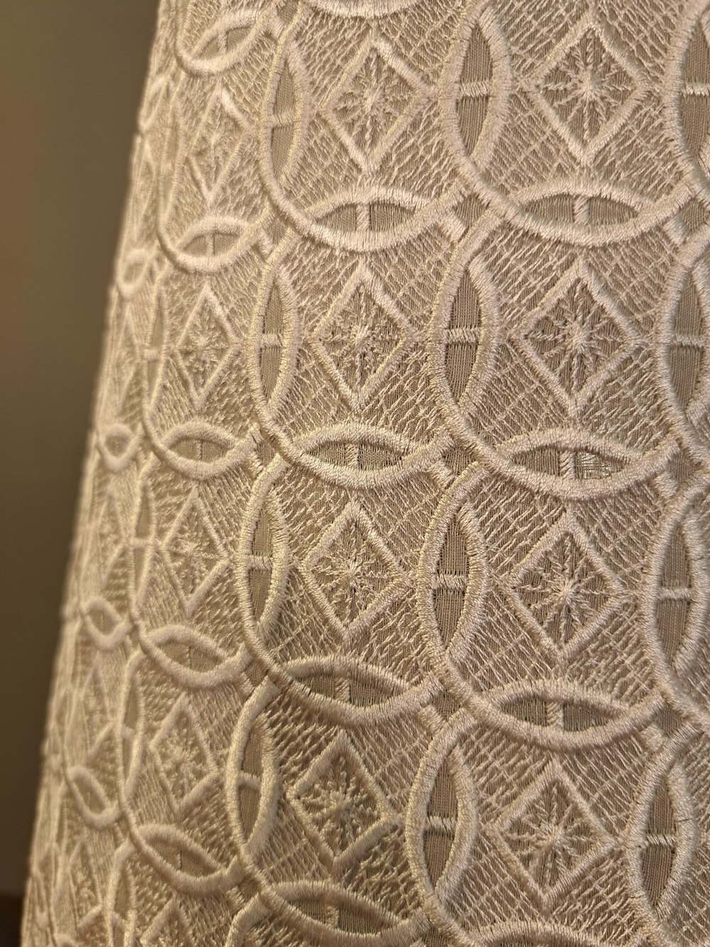 Detailaufnahme eines beigen Stoffes mit einem komplizierten geometrischen Muster aus gestickten Kreisen und Rautenformen.