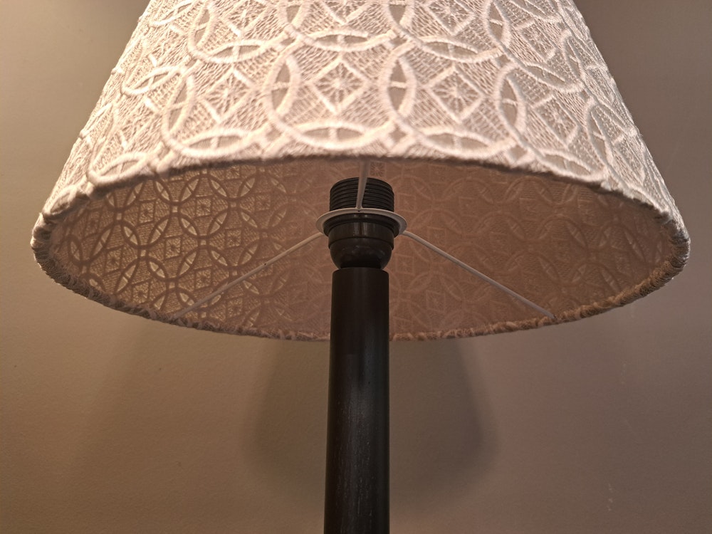 Nahaufnahme einer Lampe mit gemustertem Lampenschirm aus Stoff und dunklem Sockel vor einem schlichten Wandhintergrund.
