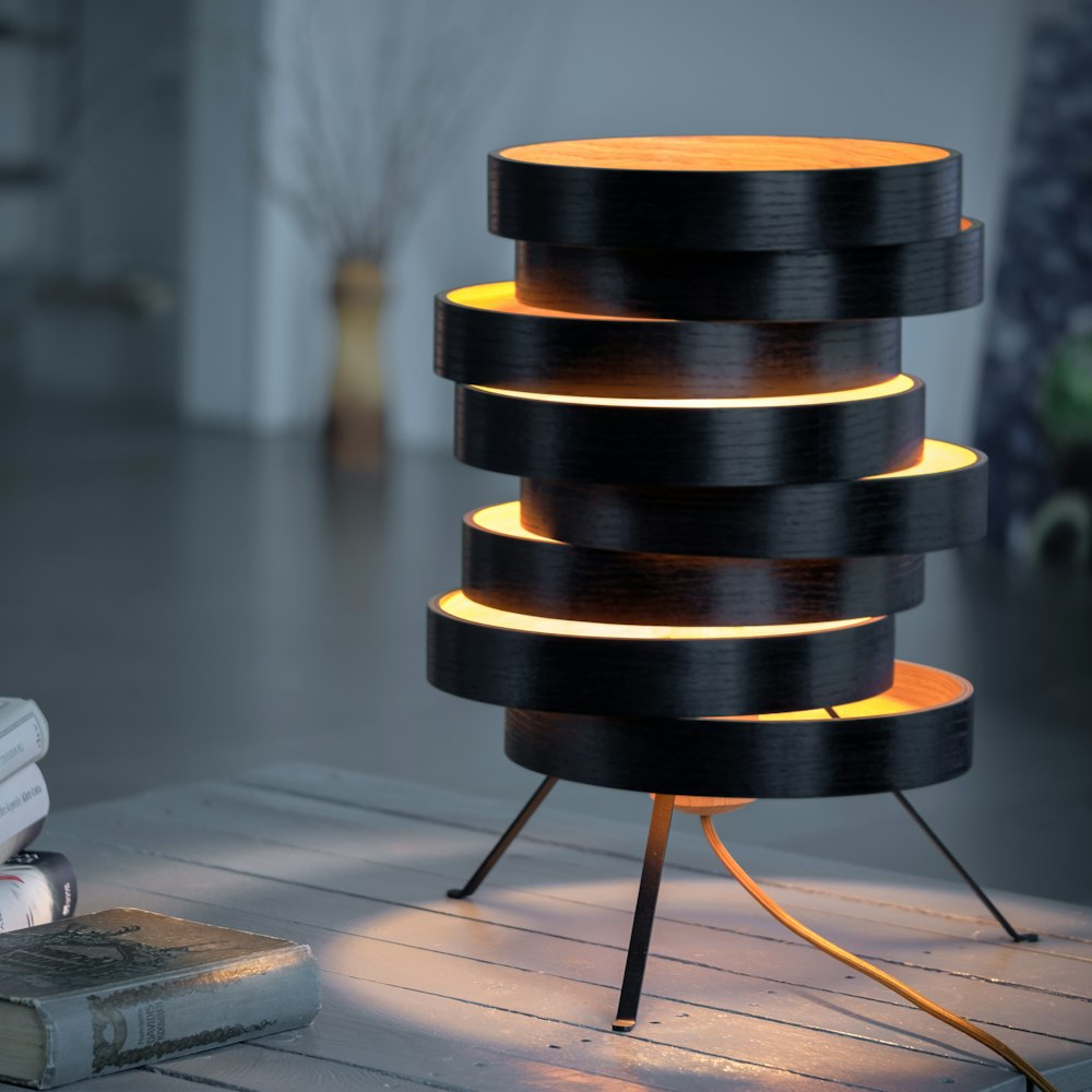 Eine moderne Lampe mit gestapelten, versetzten kreisförmigen Schichten leuchtet warm auf drei Beinen auf einem Holztisch in einem sanft beleuchteten Raum.