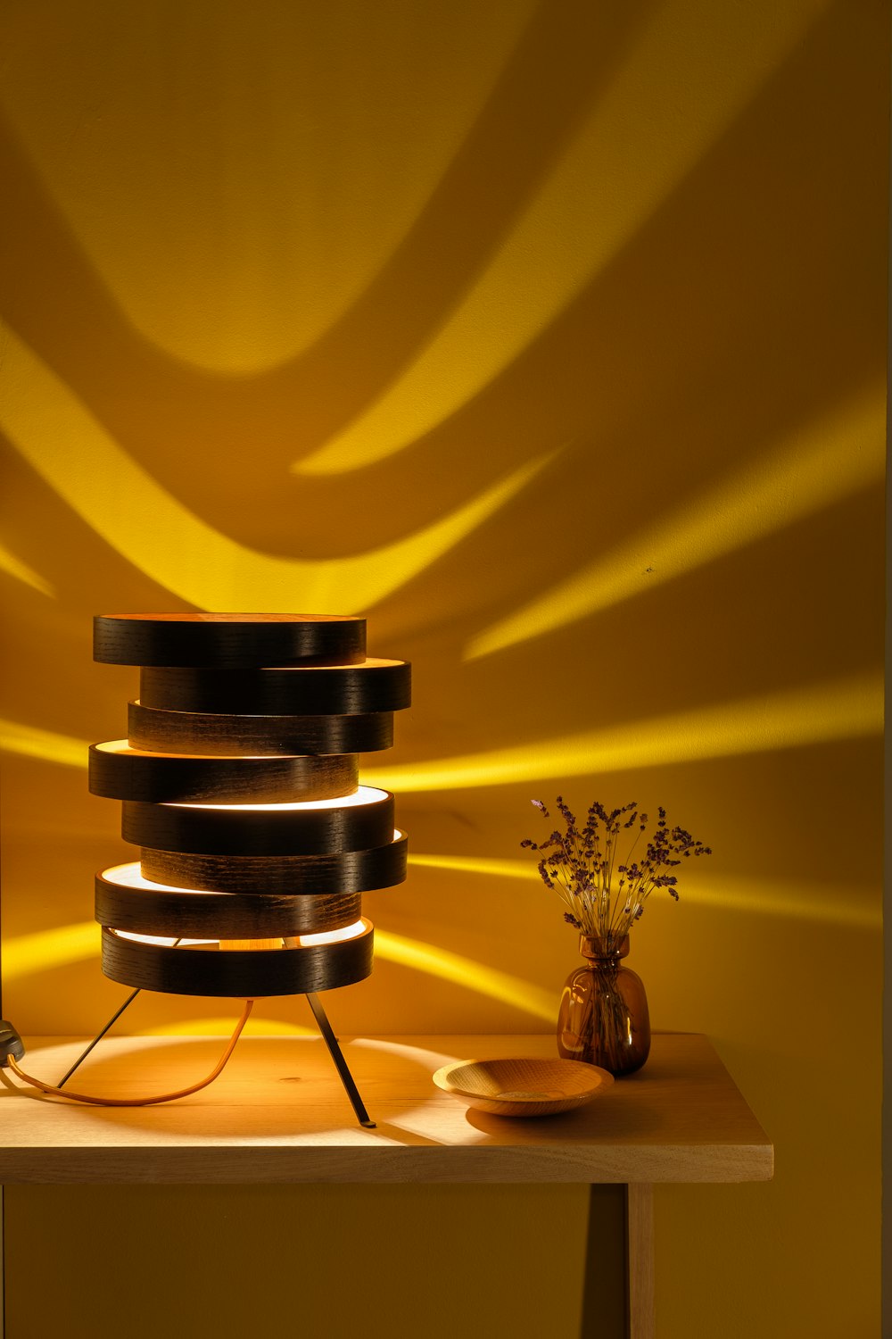 Eine moderne Holztischlampe wirft warmes Licht auf eine gelbe Wand, auf der Trockenblumen und eine Holzschale auf einem hellen Holztisch stehen.