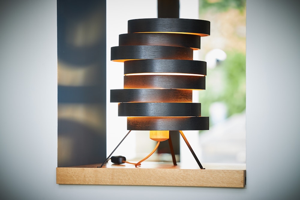 Eine moderne Tischlampe mit gestapelten runden schwarzen Schirmen leuchtet warm auf einem Holzsims am Fenster.
