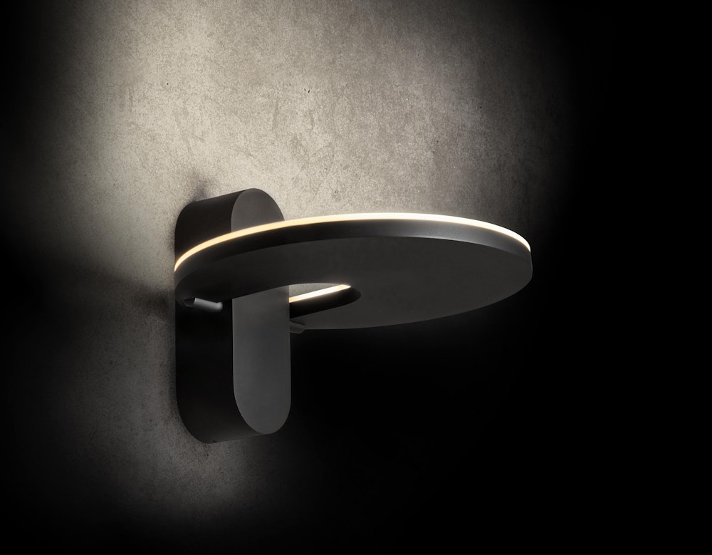 Eine kreisförmige Wandleuchte mit mattschwarzem Finish spendet weiches, diffuses Licht und ist an einer strukturierten graün Wand angebracht.