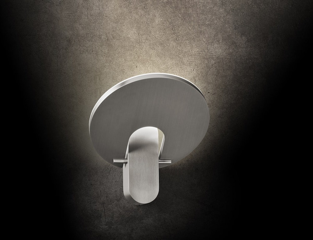 Eine moderne, runde Wandleuchte aus Metall an einer dunklen, strukturierten Wand spendet weiches, indirektes Licht, das sanft über ihr leuchtet.