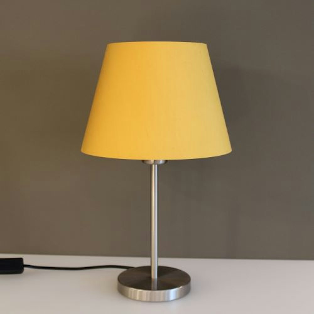 Eine Tischlampe mit gelbem Lampenschirm und Metallsockel ruht auf einer weissen Fläche vor einer graün Wand.