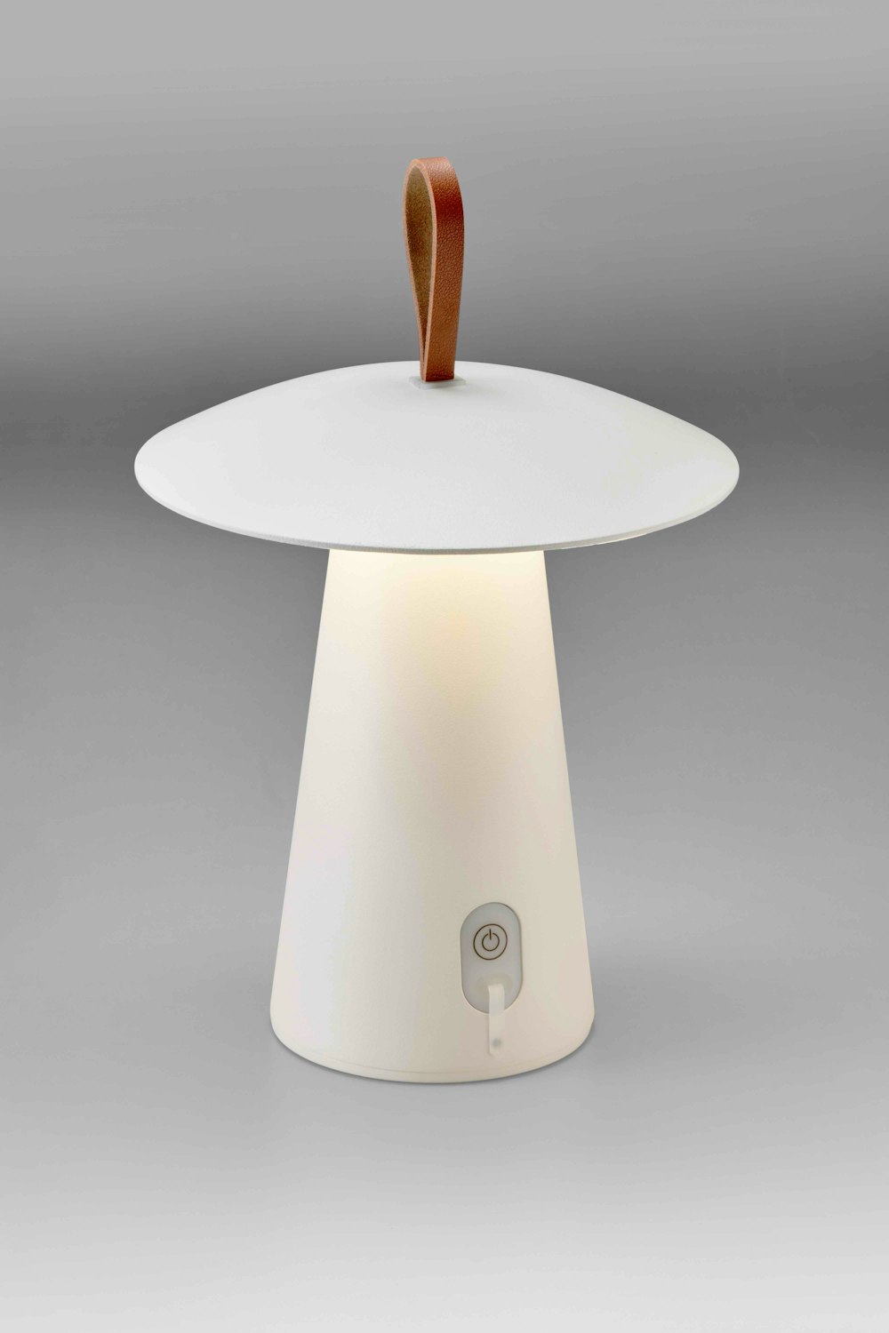 Eine moderne, weisse, pilzförmige Tischlampe mit einem Griff aus Leder und einem Einschaltknopf vor einem schlichten graün Hintergrund.