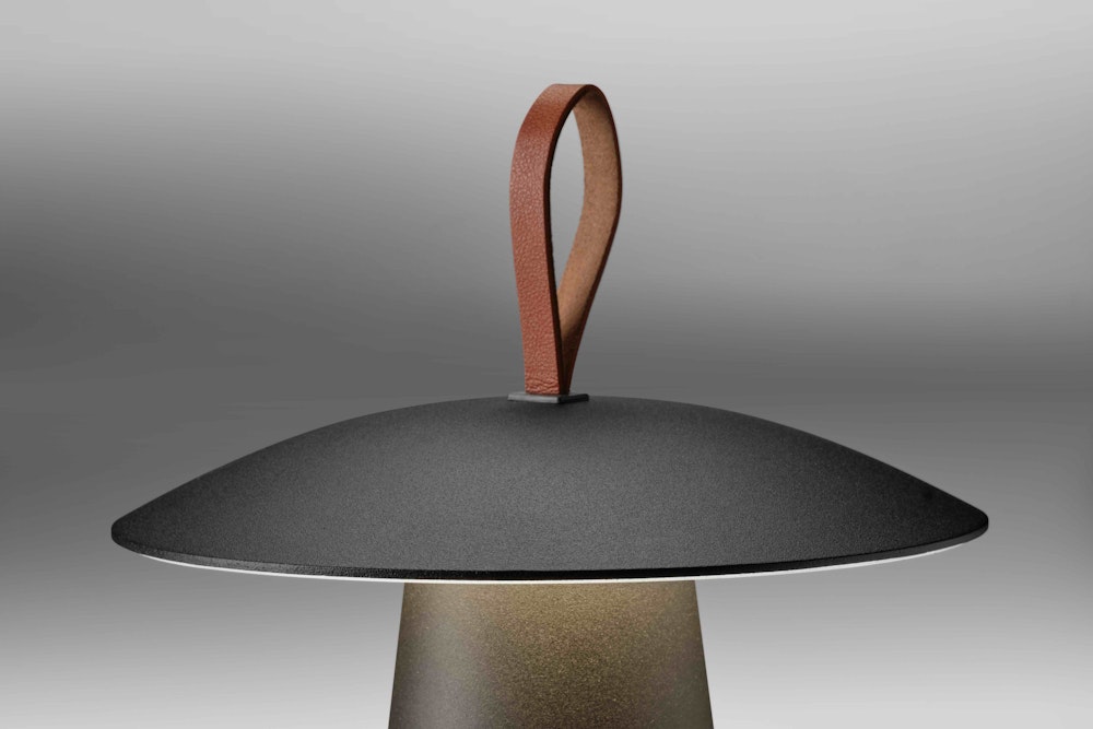 Eine moderne Lampe mit einem schwarzen, kuppelförmigen Schirm und einer braunen Lederschlaufe an der Oberseite, die sich vor einem neutralen graün Hintergrund abhebt.