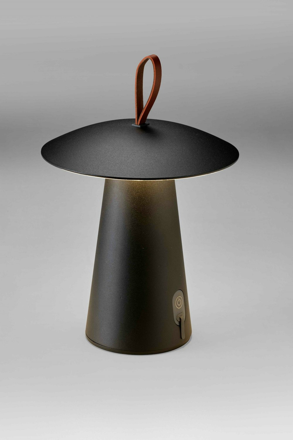 Eine moderne, kegelförmige Tischleuchte mit einem breiten schwarzen Schirm, einem Metallfuss, einem Einschaltknopf und einer braunen Lederschlaufe an der Spitze.