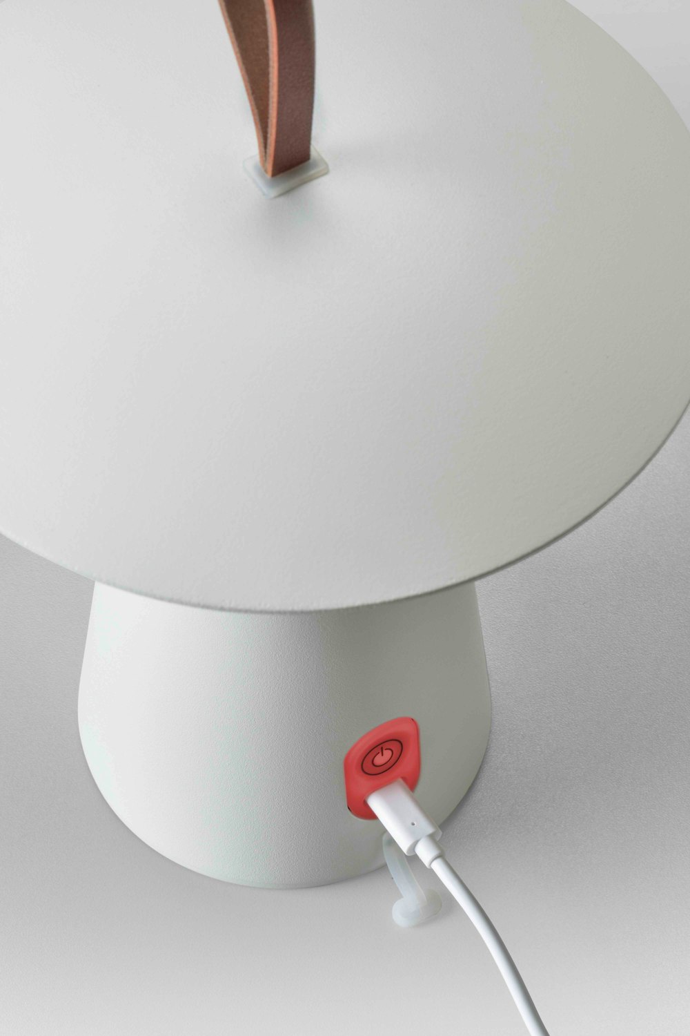 Eine weisse Lampe mit einem braunen Ledergriff, einem roten Einschaltknopf und einem an der Basis angeschlossenen Ladekabel.
