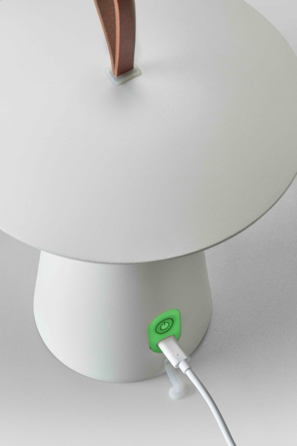 Eine weisse Lampe mit einem Ledergriff; ein Kabel ist über einen grünen Einschaltknopf mit dem Sockel verbunden.